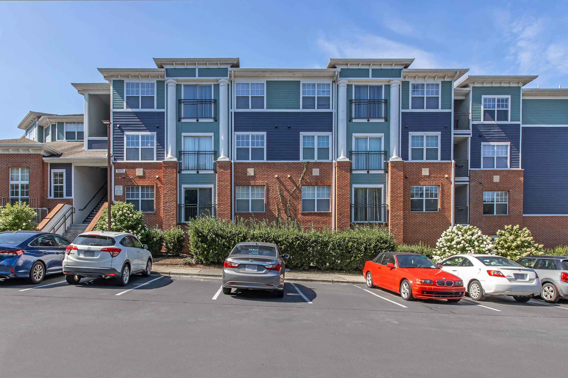 Exterior view of a multi-story apartment building featuring a mix of brick and blue siding. The building has several windows and balconies, surrounded by greenery. A parking lot in the foreground has several parked cars, including a red car and an SUV. The sky is clear with a few clouds.