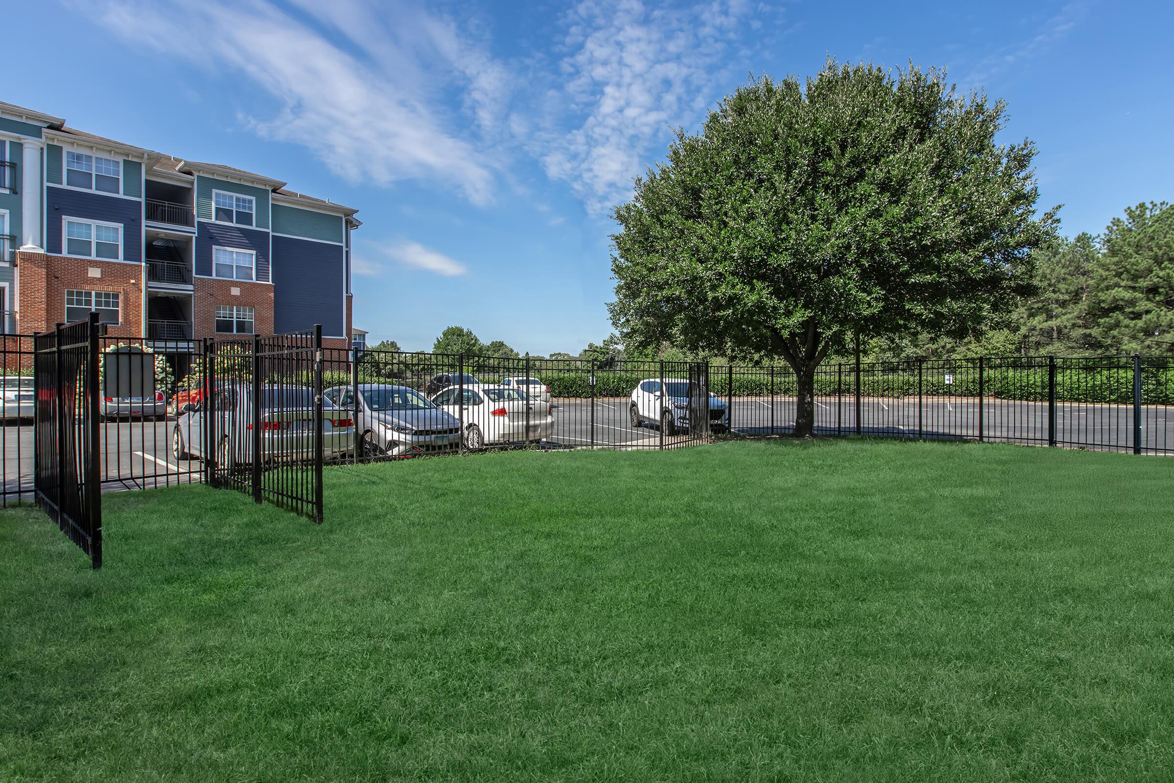 A well-maintained green lawn enclosed by a black metal fence, with several parked cars visible behind the fence. In the background, an apartment building with a mix of blue and brick exterior is partially visible, along with trees and a clear blue sky.