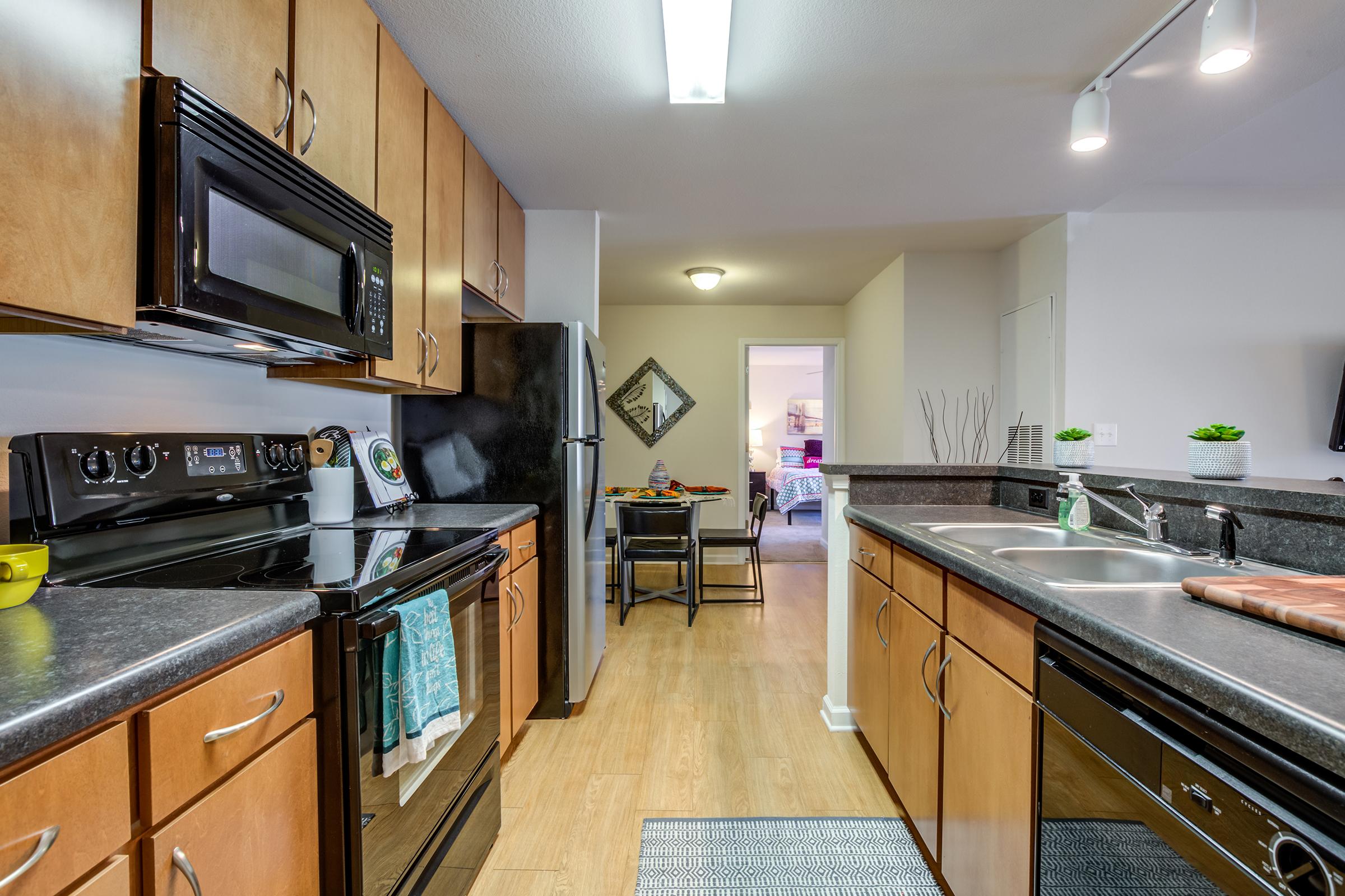 A modern kitchen featuring wooden cabinets, stainless steel appliances, and a black countertop. The kitchen includes a double sink, an oven, a microwave, and a refrigerator. A small dining area is visible in the background, adding a cozy touch to the space. Natural light brightens the area.