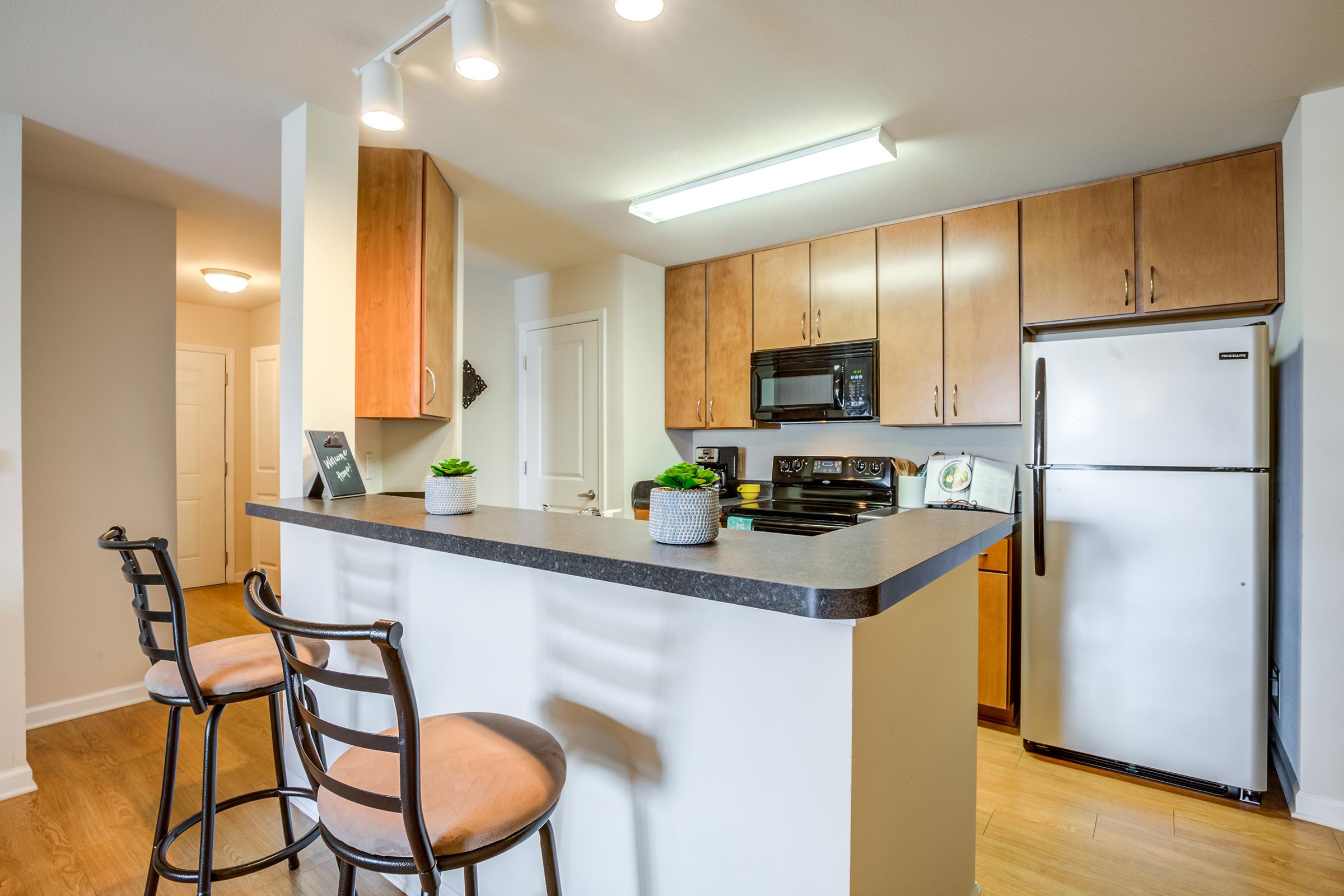 A modern kitchen featuring wooden cabinetry, stainless steel appliances, and a sleek countertop. Two bar stools are positioned at the kitchen island, and the space is well-lit with overhead lighting. A doorway leads to another room, adding depth to the layout. Small decorative plants are placed on the counter for a touch of greenery.