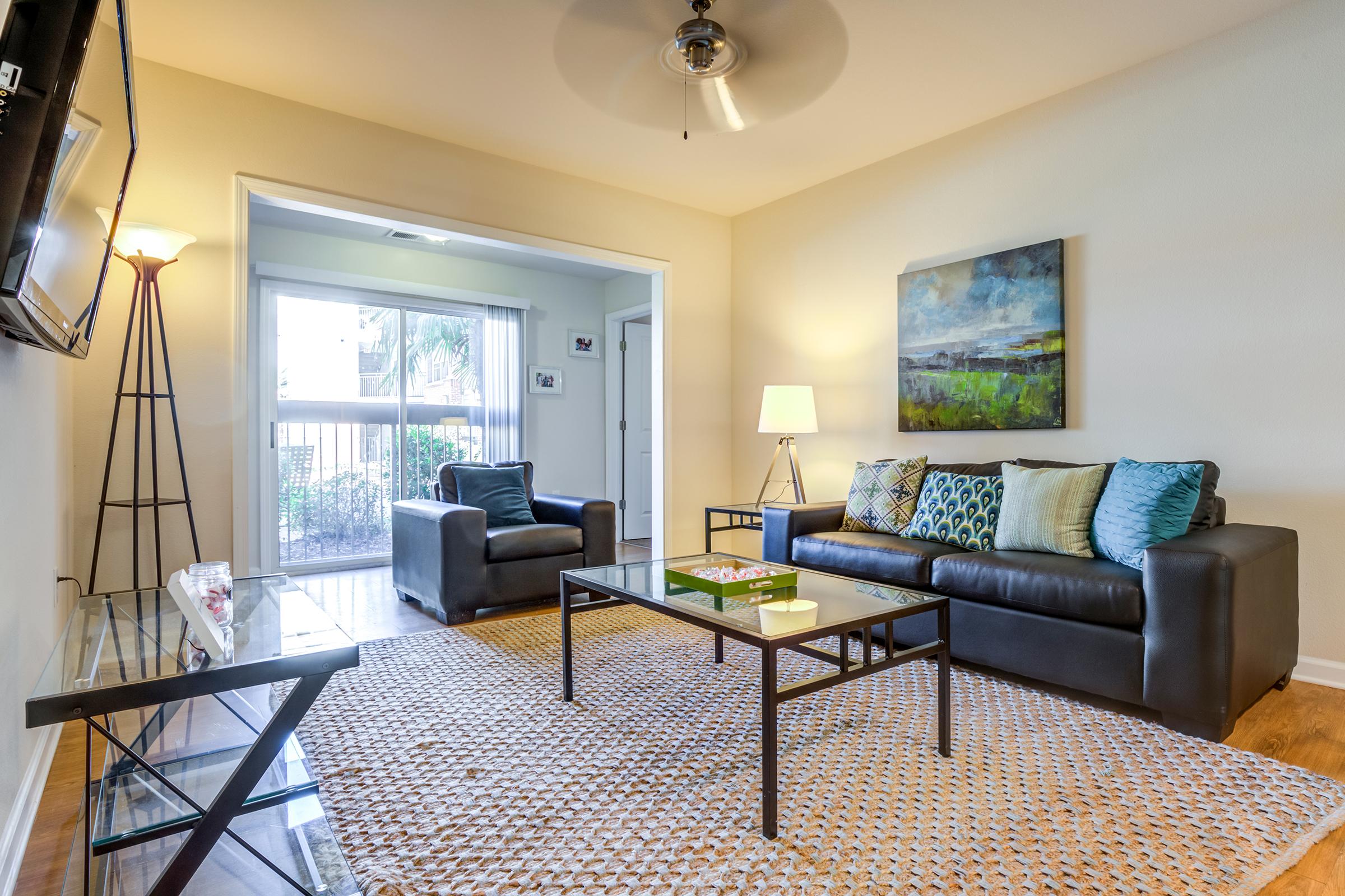 Cozy living room featuring a black leather sofa with decorative cushions, a glass coffee table, and two armchairs. A floor lamp and wall-mounted TV are visible. Natural light comes through a nearby window, enhancing the warm atmosphere. A textured area rug adds comfort to the space.