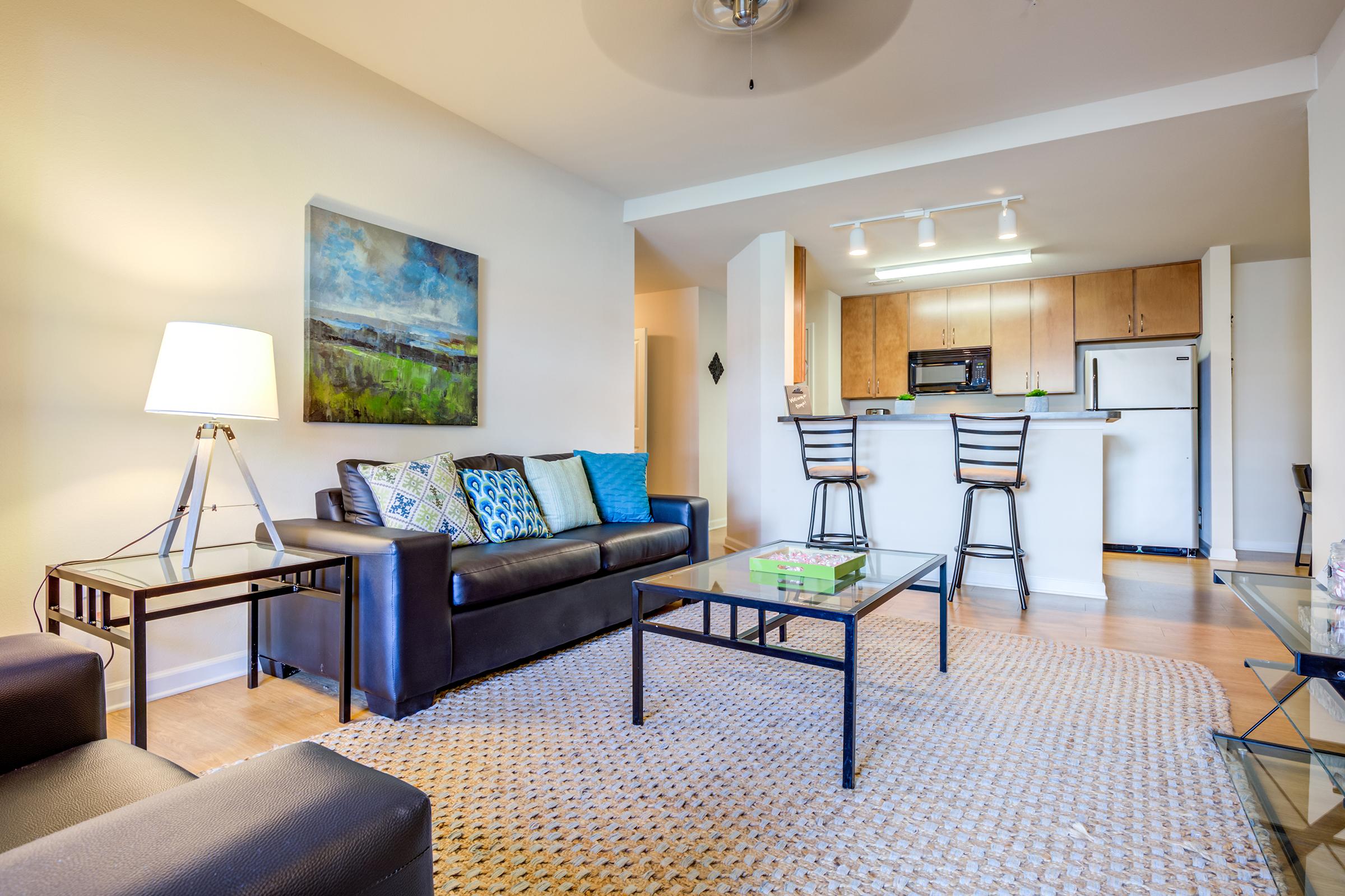Modern living room with a dark leather sofa adorned with decorative pillows, a glass coffee table, and a light-colored area rug. A lamp provides warm lighting, and a colorful painting hangs on the wall. The open kitchen area features wooden cabinets, bar stools, and modern appliances. Bright and inviting atmosphere.