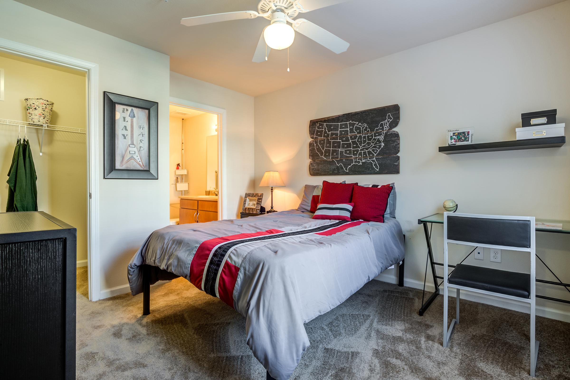 A cozy bedroom featuring a queen-sized bed with a gray and red bedspread, a ceiling fan, and a small desk with a chair. There's a framed map on the wall and a closet in the corner. The room has neutral-colored walls and carpet, offering a warm and inviting atmosphere. A bathroom is visible through an open door.