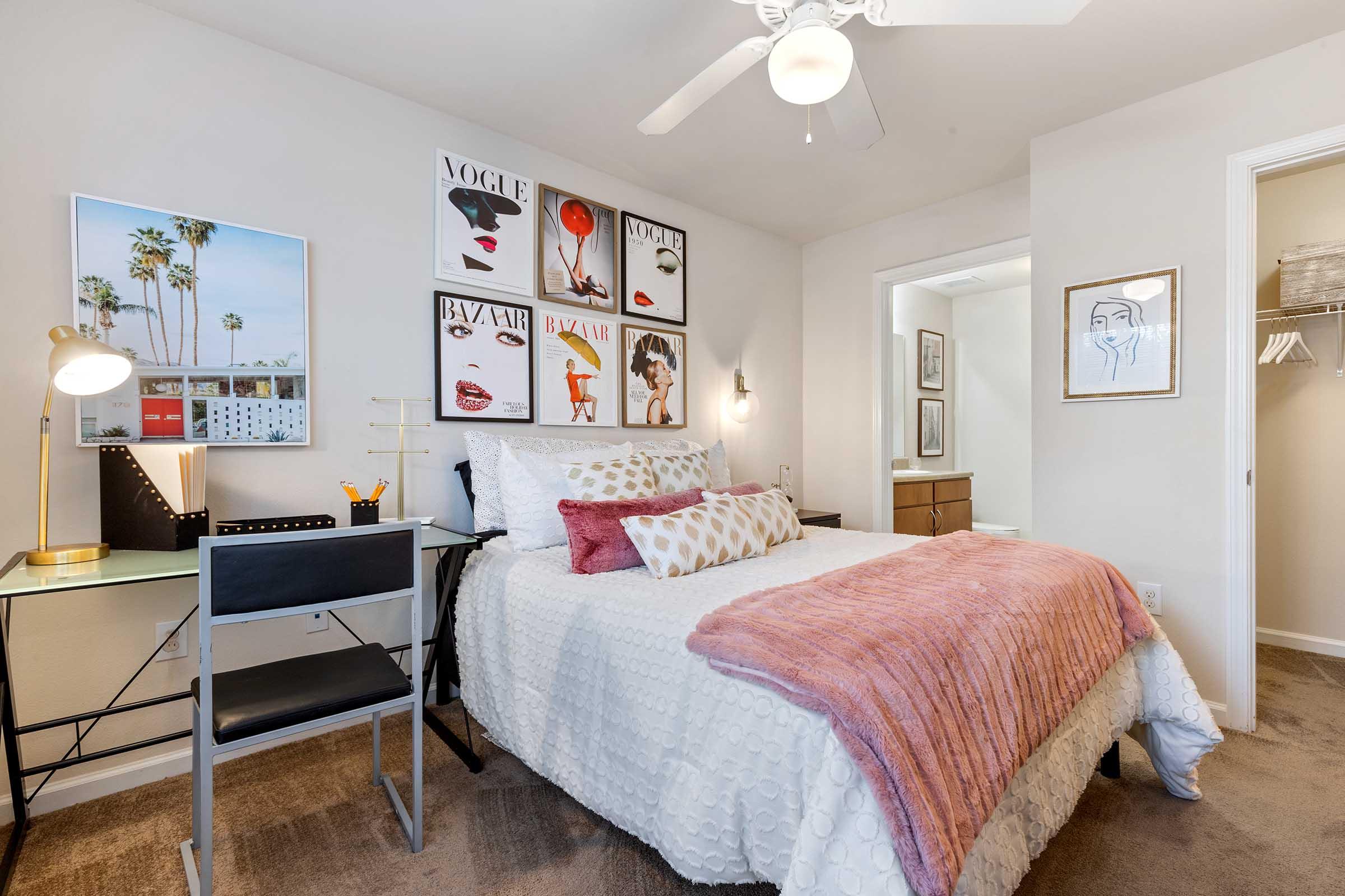 Cozy bedroom featuring a white quilted bed with pink throw blanket, a desk with a lamp and chair, and stylish wall art. The room has a ceiling fan and beige walls, creating a warm atmosphere. A doorway leads to an adjoining space with additional decor.
