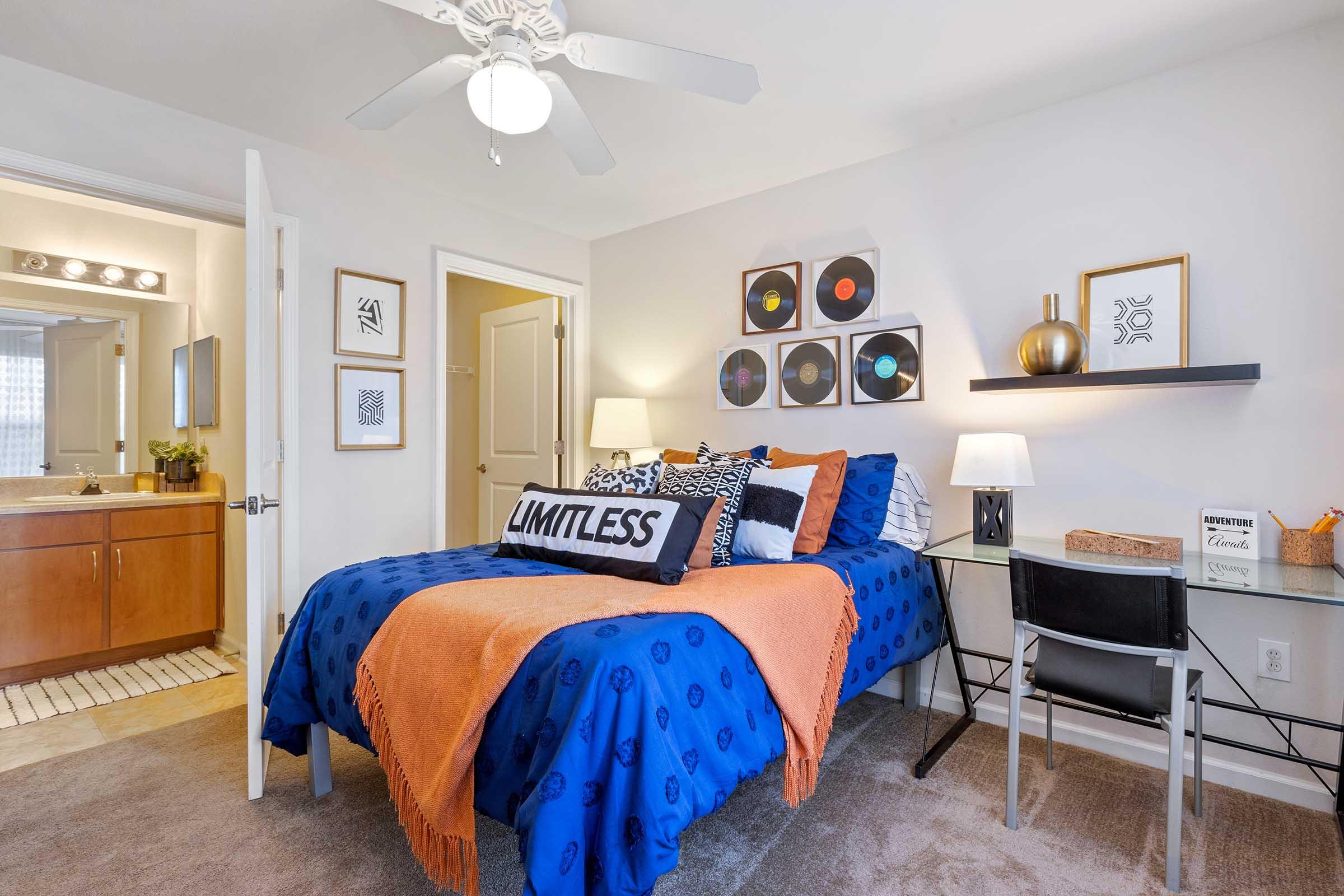 Cozy bedroom featuring a queen-sized bed with a blue and orange color scheme, decorative pillows, and a "LIMITLESS" throw pillow. A desk with a chair is positioned beside the bed, and a wall displays framed art and vinyl records. A bathroom is visible through an open door, adding to the room's inviting atmosphere.