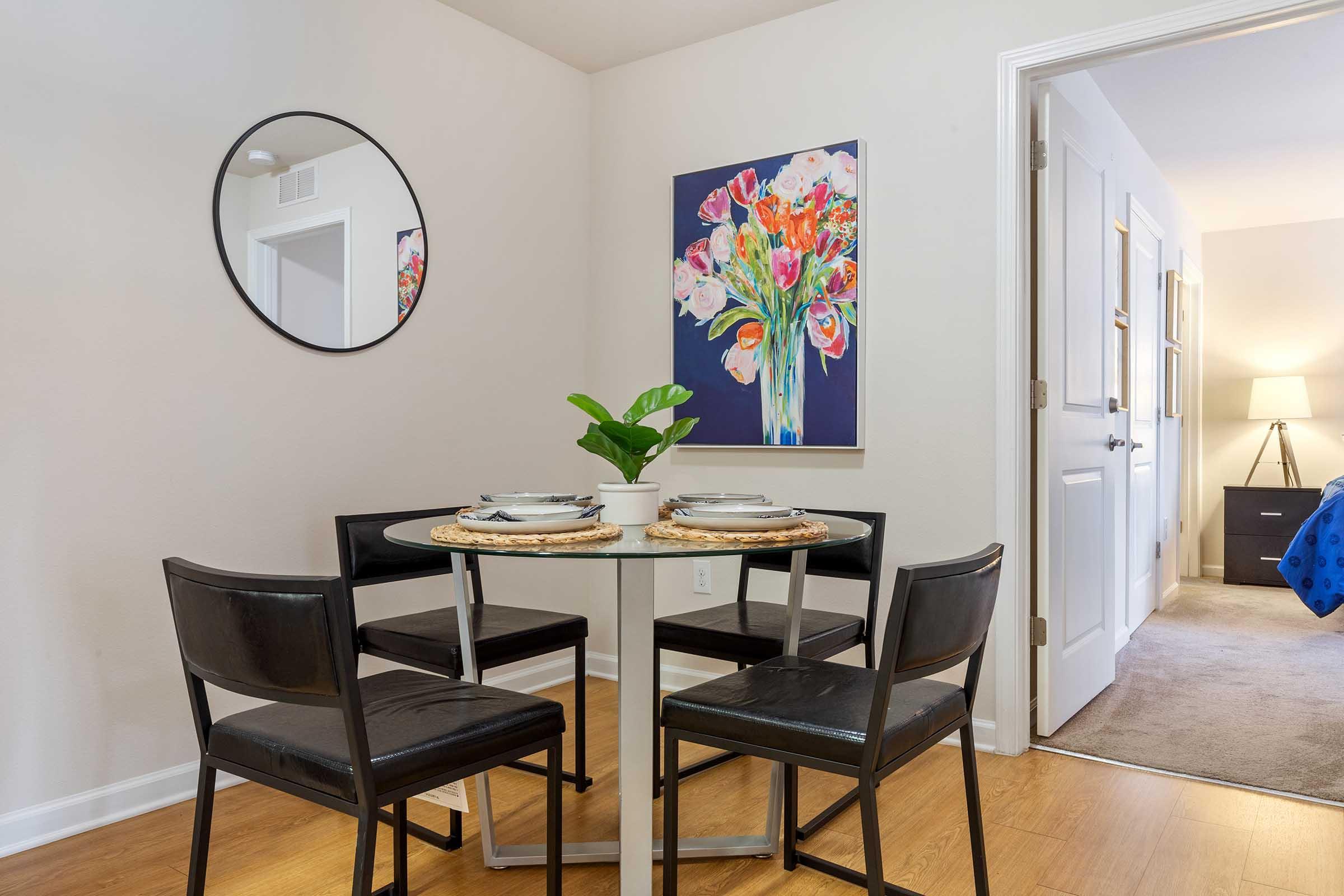 A modern dining area featuring a round glass table surrounded by four black chairs. A small potted plant sits on the table. A vibrant floral painting hangs on the wall, and a round mirror reflects the space. A doorway leads to another room, adding to the cozy atmosphere of the setting.