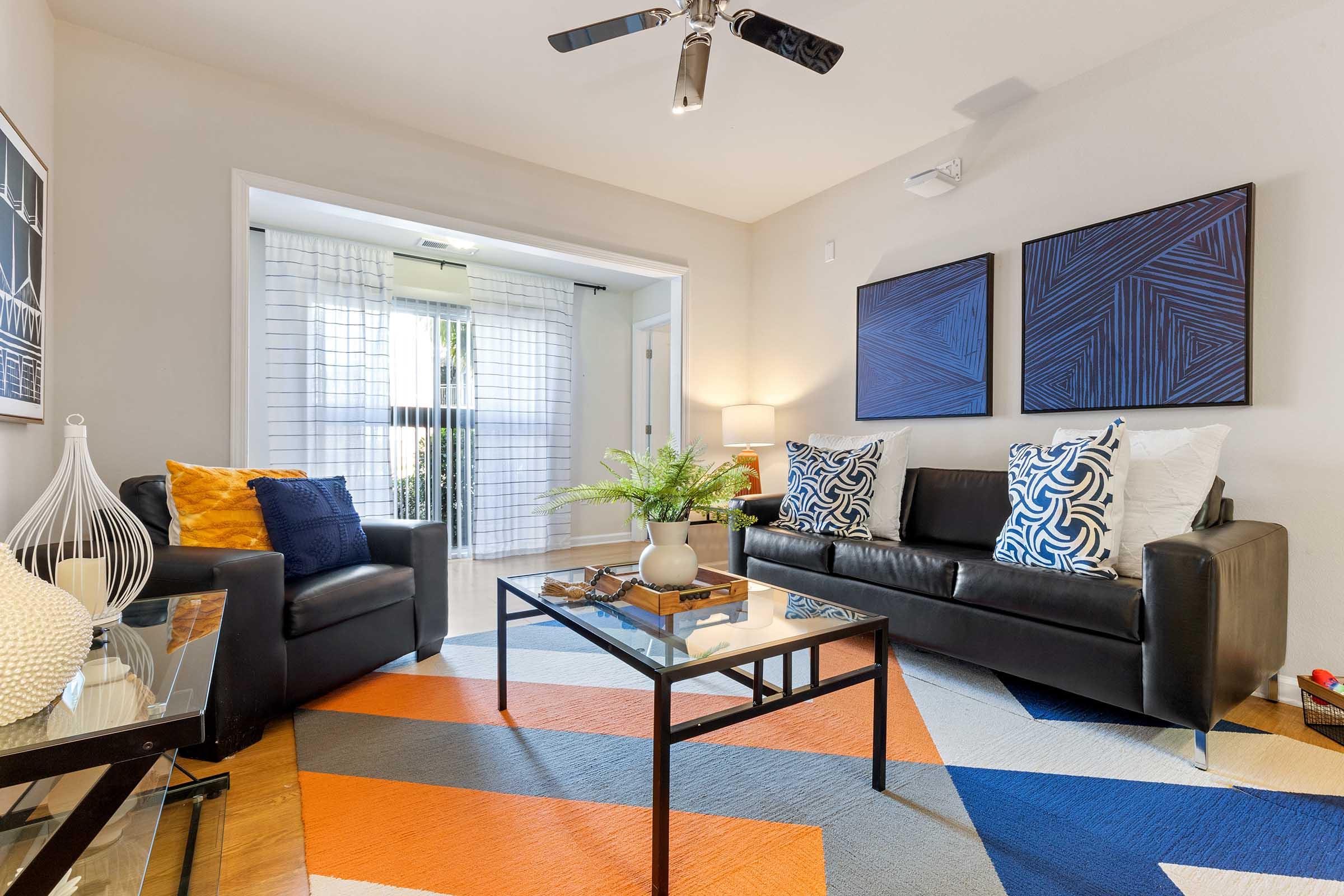 A stylish living room featuring a black leather sofa with decorative pillows, a glass coffee table, and a vibrant area rug with orange, blue, and cream patterns. There are artistic wall pieces, a small plant on the coffee table, and a cozy armchair, creating a welcoming atmosphere. Bright natural light enters through large windows.
