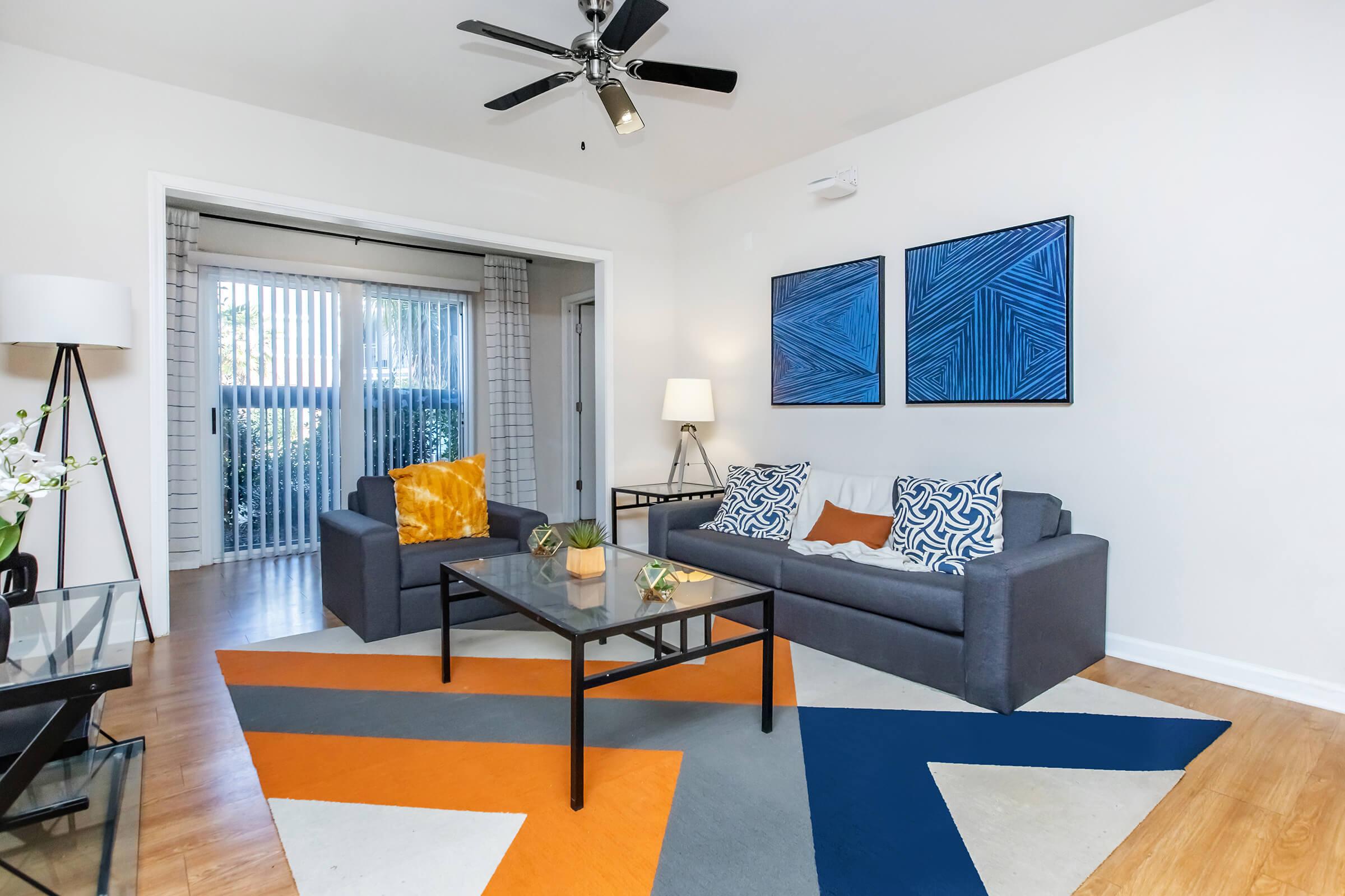 Modern living room featuring a gray sofa with colorful throw pillows, a glass coffee table, and a patterned area rug. Two abstract blue artworks hang on the wall, and there is a floor lamp and a plant on the table. A sliding glass door provides natural light and leads to an outdoor area.