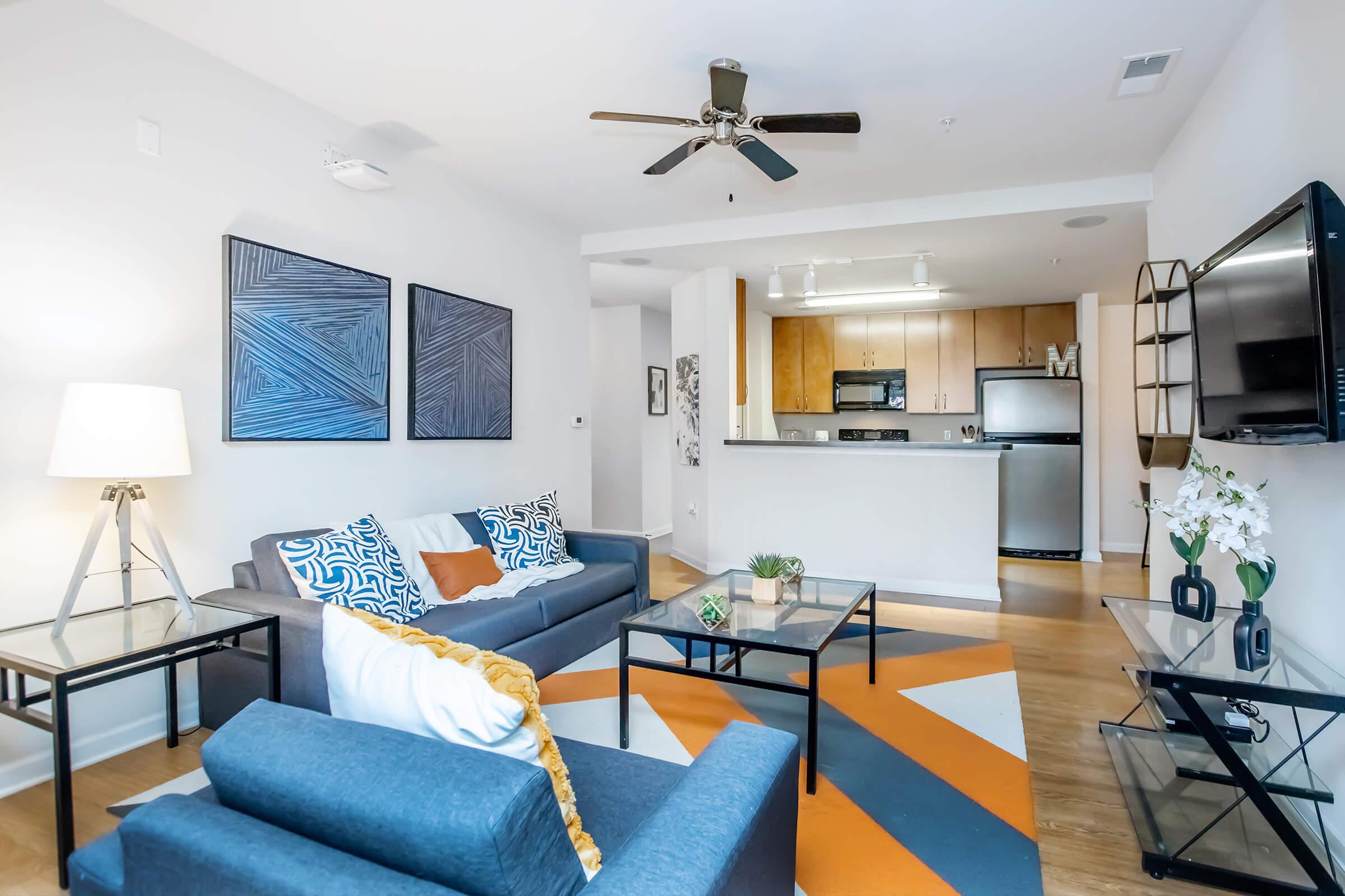 Living room featuring a gray sofa with colorful throw pillows, a matching blue armchair, and a geometric patterned area rug. There are two abstract wall art pieces, a floor lamp, a coffee table, and a TV mounted on the wall. The kitchen is visible in the background, with wooden cabinetry and stainless steel appliances.