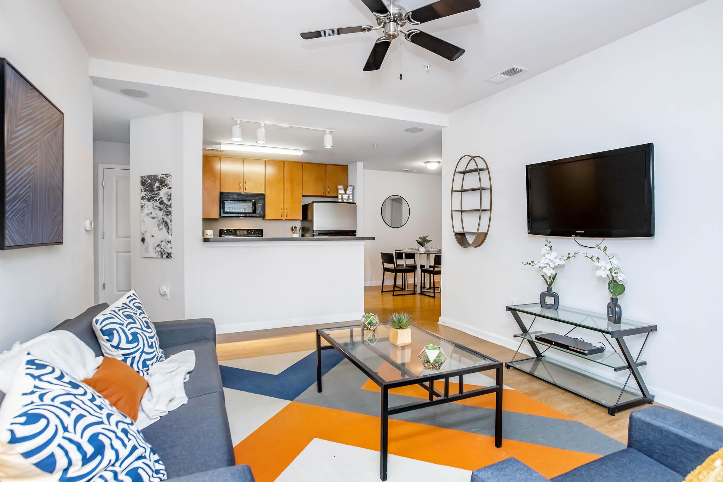 A modern living room featuring a gray sofa with colorful cushions, a geometric patterned area rug, a glass coffee table with decorative plants, and a television mounted on the wall. In the background, a kitchen area with wooden cabinets and a dining table is visible. The space is well-lit and stylishly decorated.