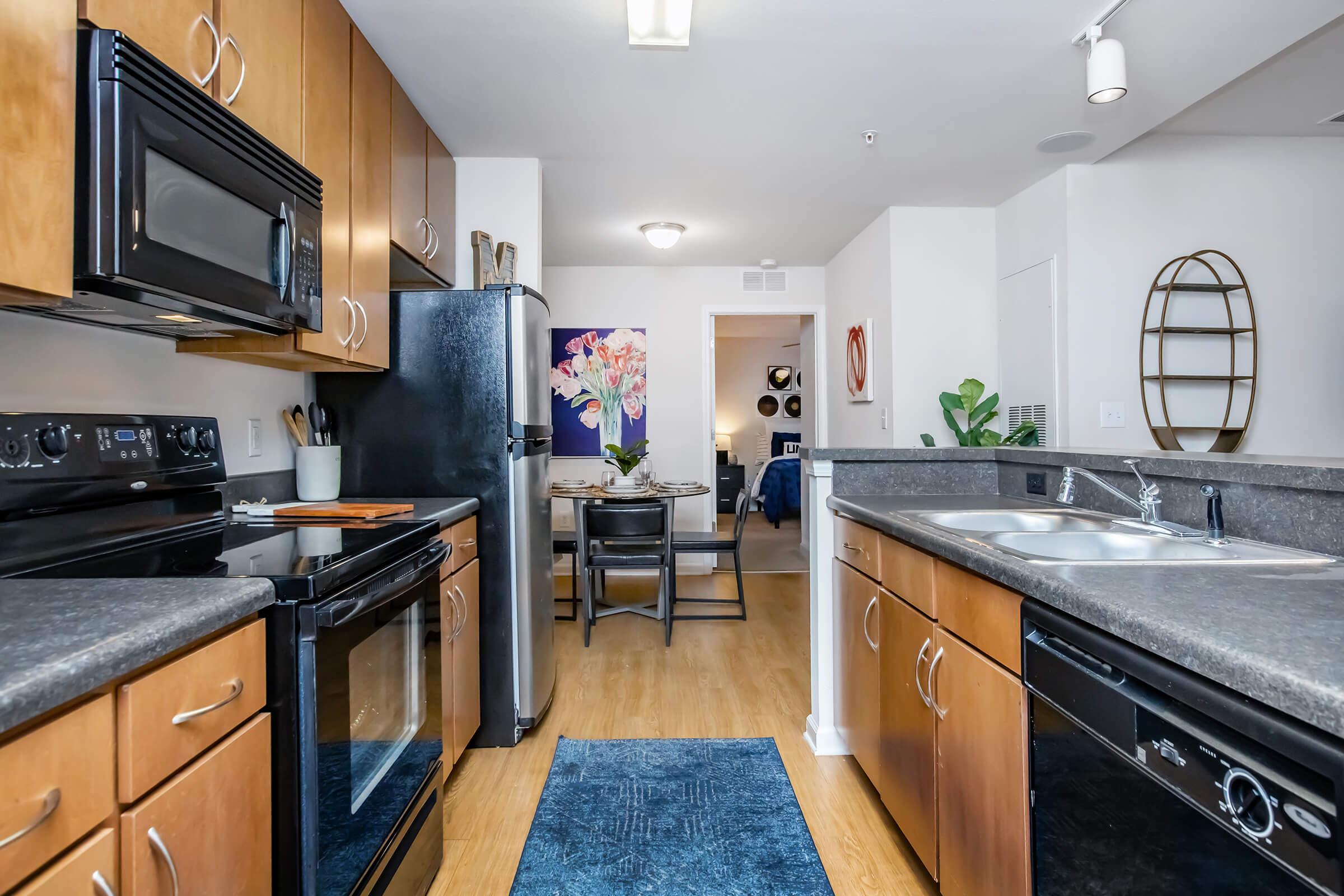 Modern kitchen featuring dark wood cabinetry, stainless steel and black appliances, and a double sink. A blue area rug lies on the wooden floor. In the background, a dining area is visible with a round table, chairs, and decorative wall art. Natural light brightens the space.