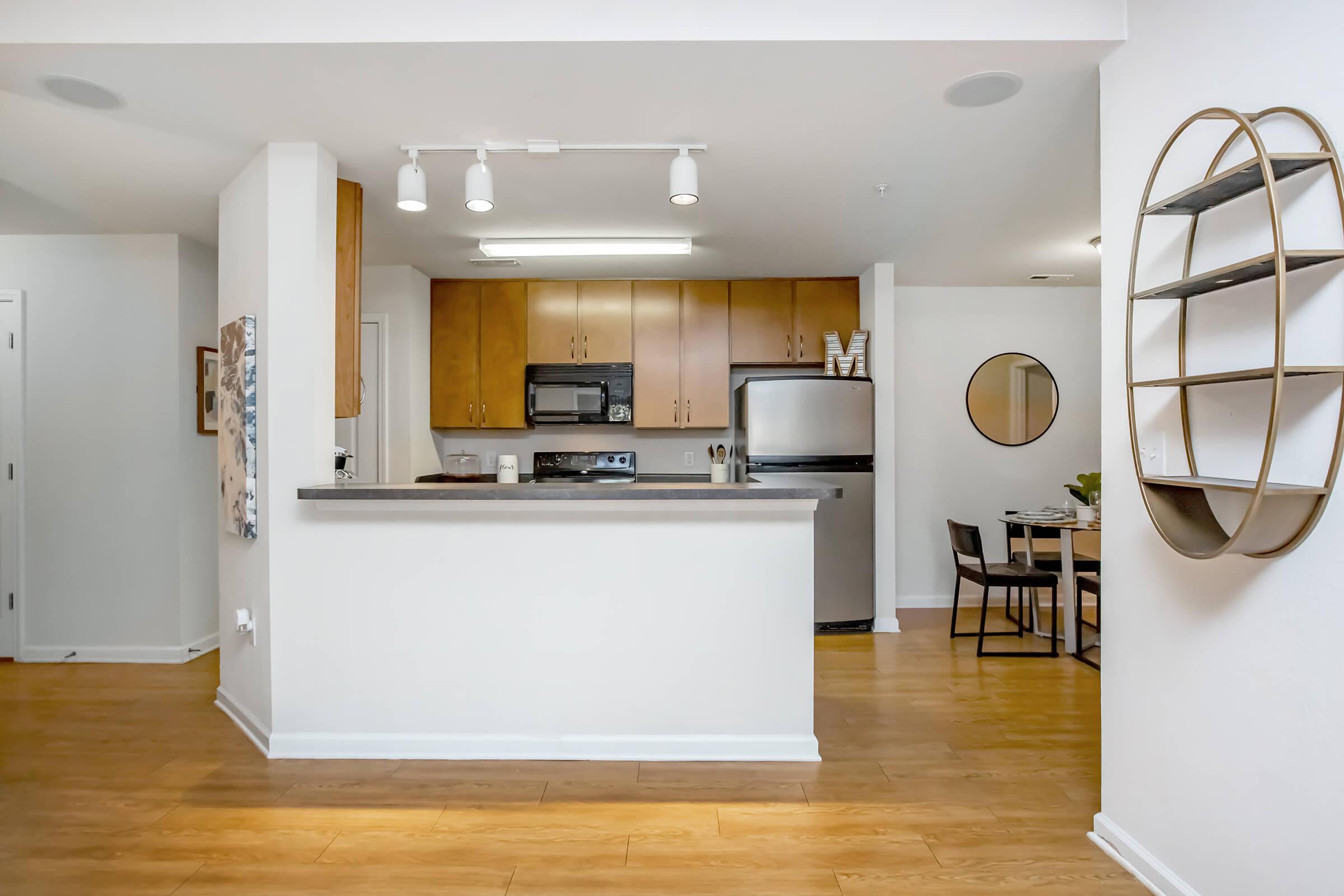 Modern kitchen and dining area featuring wood cabinetry, stainless steel appliances, and a circular mirror on the wall. The kitchen bar separates the space, with a dining table set for four visible in the background. Light-colored wood flooring enhances the open and inviting atmosphere.
