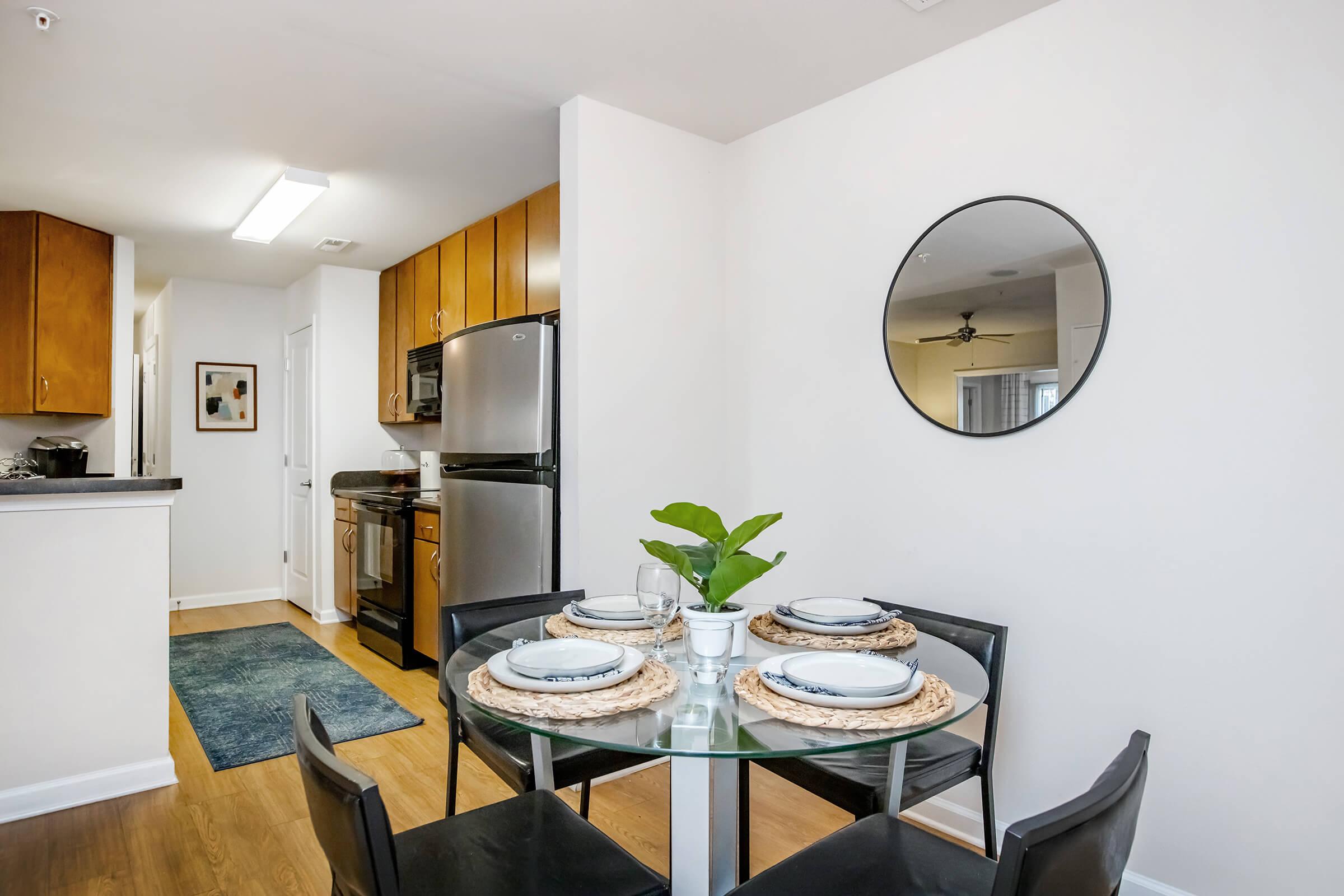 A small dining area featuring a round glass table with black chairs, set with white plates and woven placemats. A circular mirror hangs on the wall, reflecting the light. In the background, there is a modern kitchen with wooden cabinets and stainless steel appliances. A green plant adds a touch of nature to the space.
