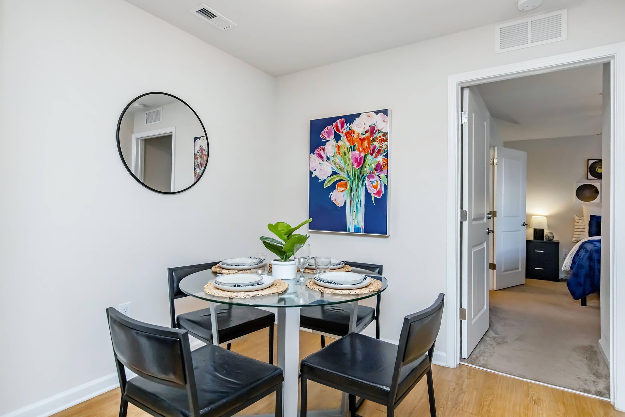 A modern dining area featuring a round glass table set for four with plates and cutlery. Black chairs surround the table. A colorful floral painting hangs on the wall, and a round mirror is mounted nearby. In the background, a doorway leads to a well-decorated bedroom.