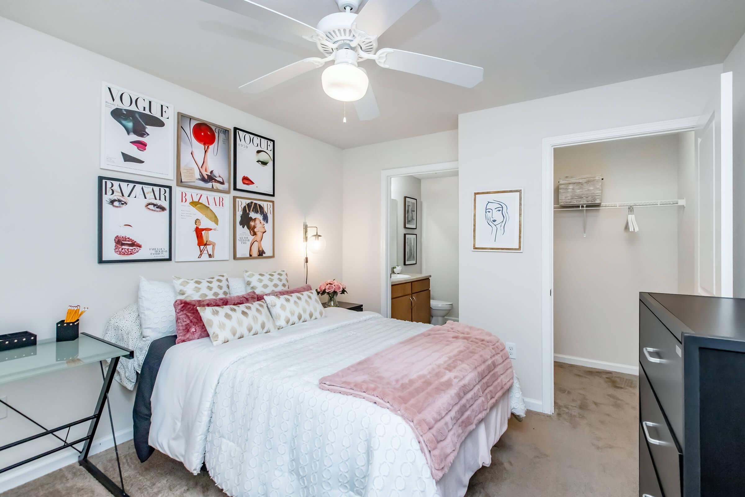 A cozy bedroom featuring a queen-sized bed with decorative pillows, a light pink throw blanket, and a stylish ceiling fan. The wall displays fashion-themed art prints. A small desk is next to the bed, and there’s an ensuite bathroom visible through the open door, along with a closet.