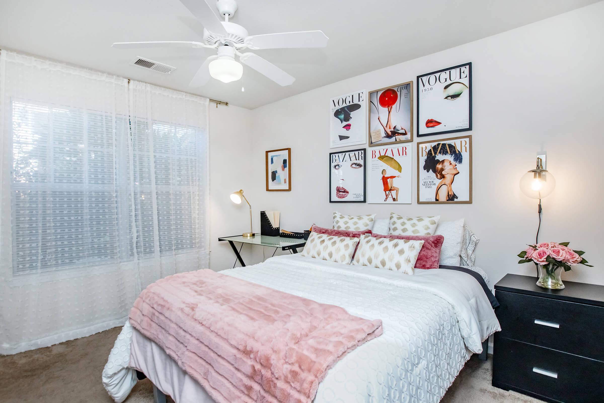 A cozy bedroom featuring a queen-sized bed with a pink and white throw blanket, decorative pillows, and stylish wall art including magazine covers. A desk lamp and a small desk are nearby, and there's a bouquet of pink flowers on a nightstand. Natural light filters through sheer curtains.