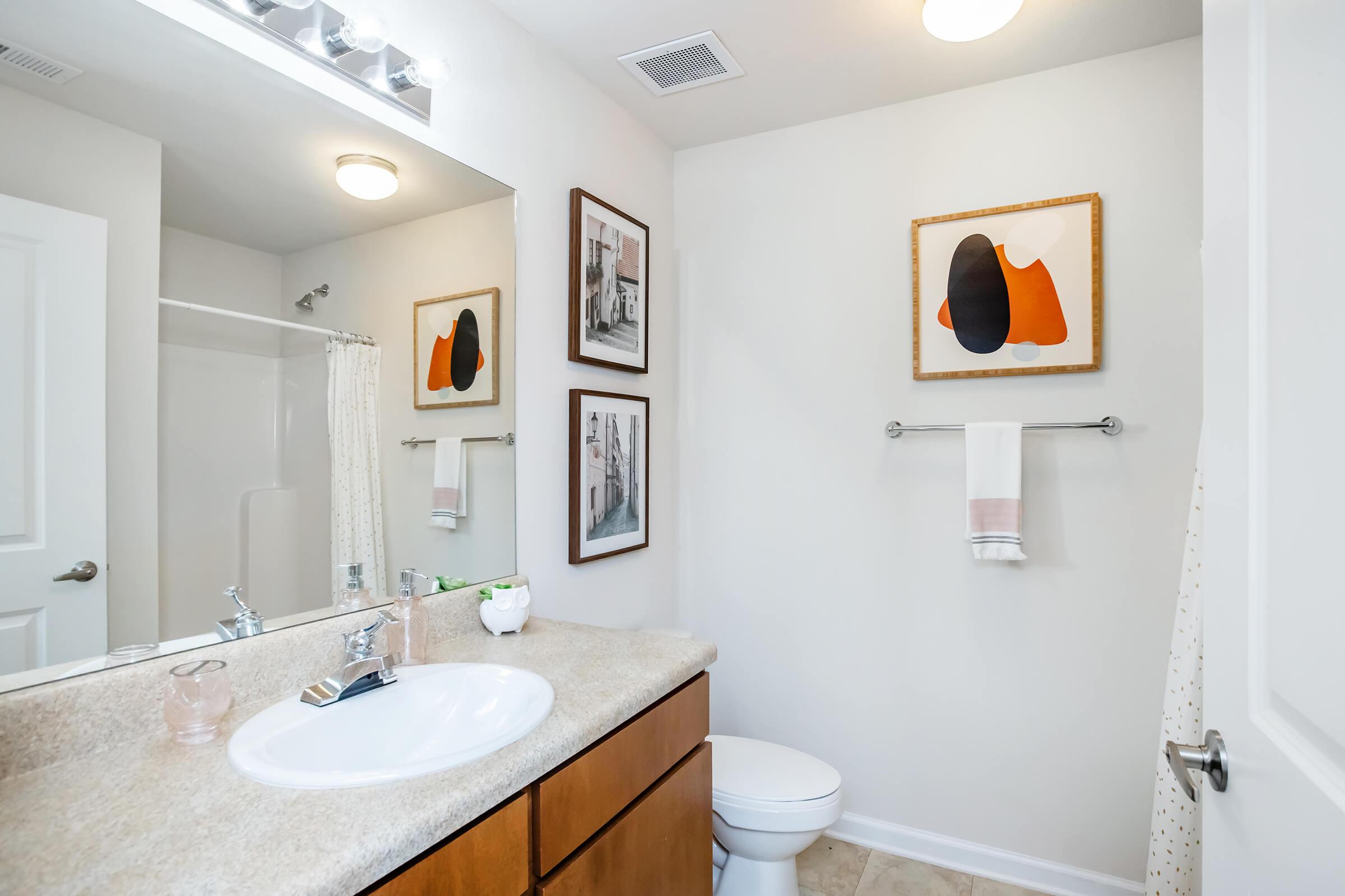 A modern bathroom featuring a sink with a wooden cabinet, a shower stall, and a toilet. The walls are painted white, and there are framed artworks on the wall above the sink. A towel hangs on a silver rack, and decorative items are placed on the countertop. Natural light illuminates the space.