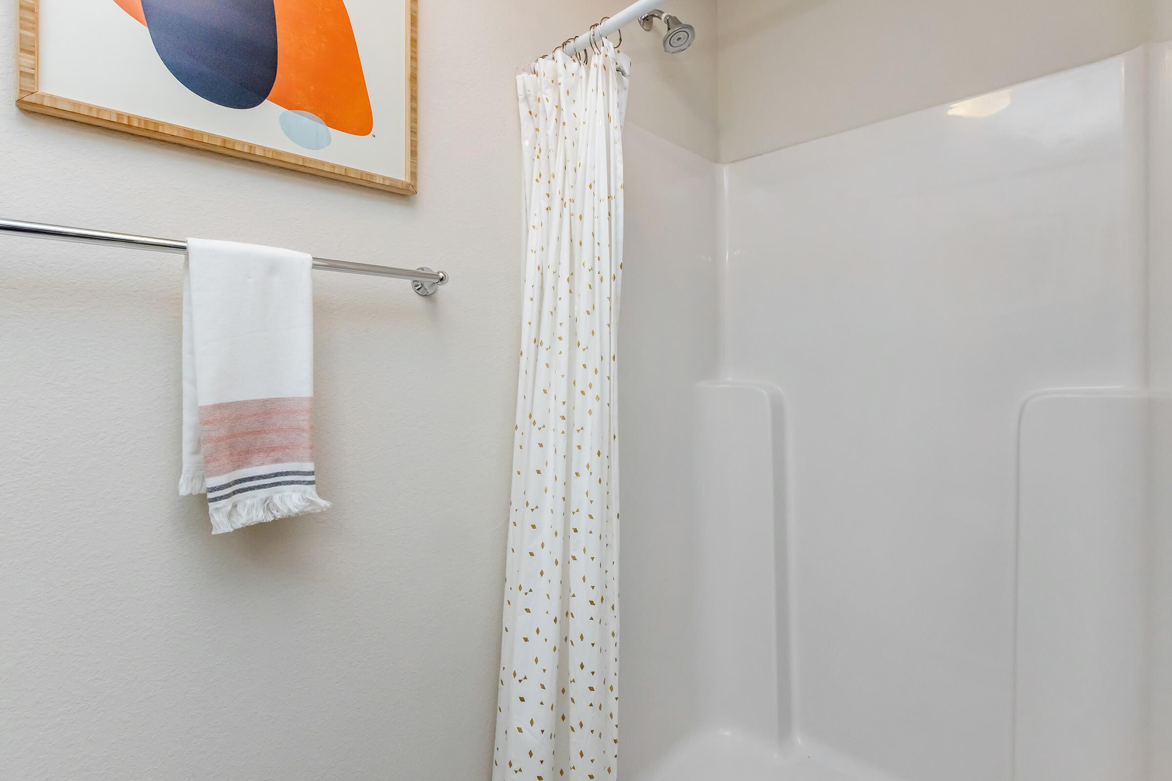 A bright, clean bathroom featuring a white shower stall with a decorative curtain. A towel is neatly hanging on a silver rack beside the shower. Above, a framed artwork with abstract shapes in orange and blue adds a splash of color to the minimalist space.