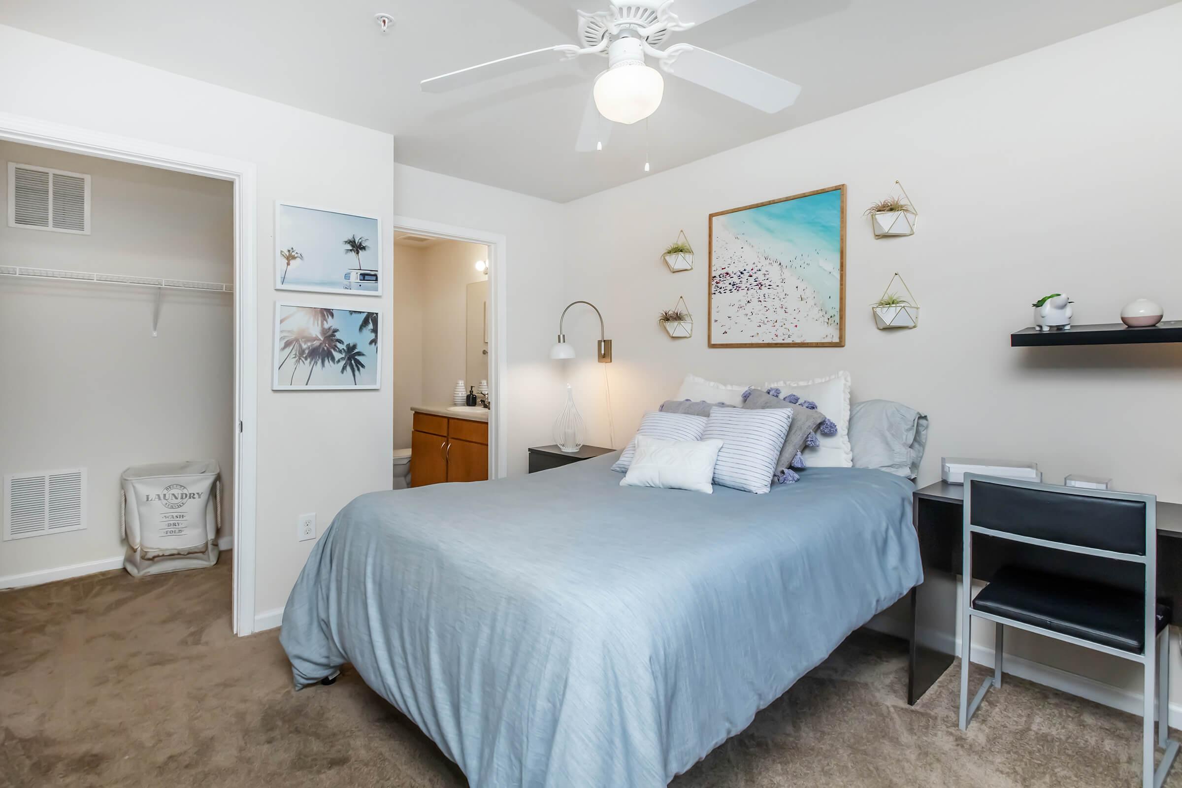 A cozy bedroom featuring a queen-sized bed with blue bedding and decorative pillows. There's a small desk with a chair, a ceiling fan, framed artwork on the wall, and plants in wall-mounted holders. A closet is visible, and a bathroom door is seen in the background. The room has a warm, inviting ambiance.