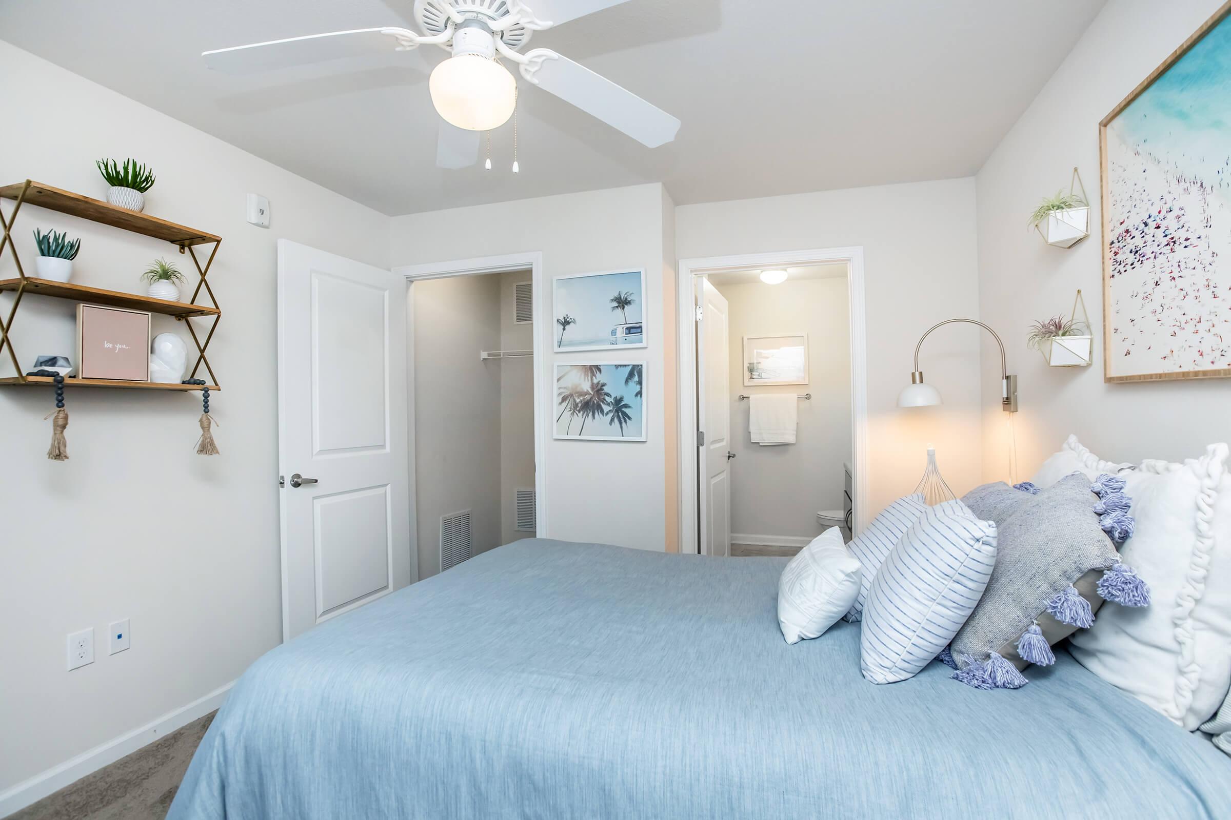 A cozy bedroom featuring a light blue bedspread with decorative pillows, a ceiling fan, and a wall-mounted reading lamp. There's a wooden shelf with potted plants and artwork on the walls, along with a closet door and a bathroom entrance in the background, all contributing to a warm, inviting atmosphere.