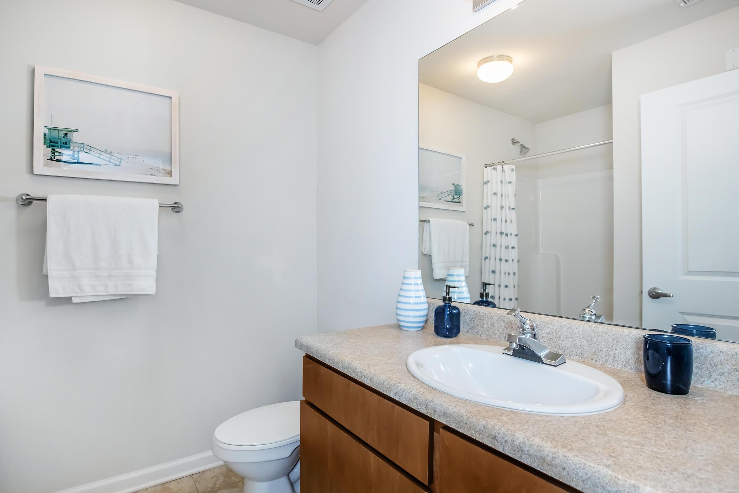 A modern, well-lit bathroom featuring a light-colored countertop with a sink, a large mirror, and wooden cabinets. There are white towels hanging on a rack, a shower stall with a curtain, and decorative items like a blue vase. A framed beach photo adds a touch of decor to the wall.