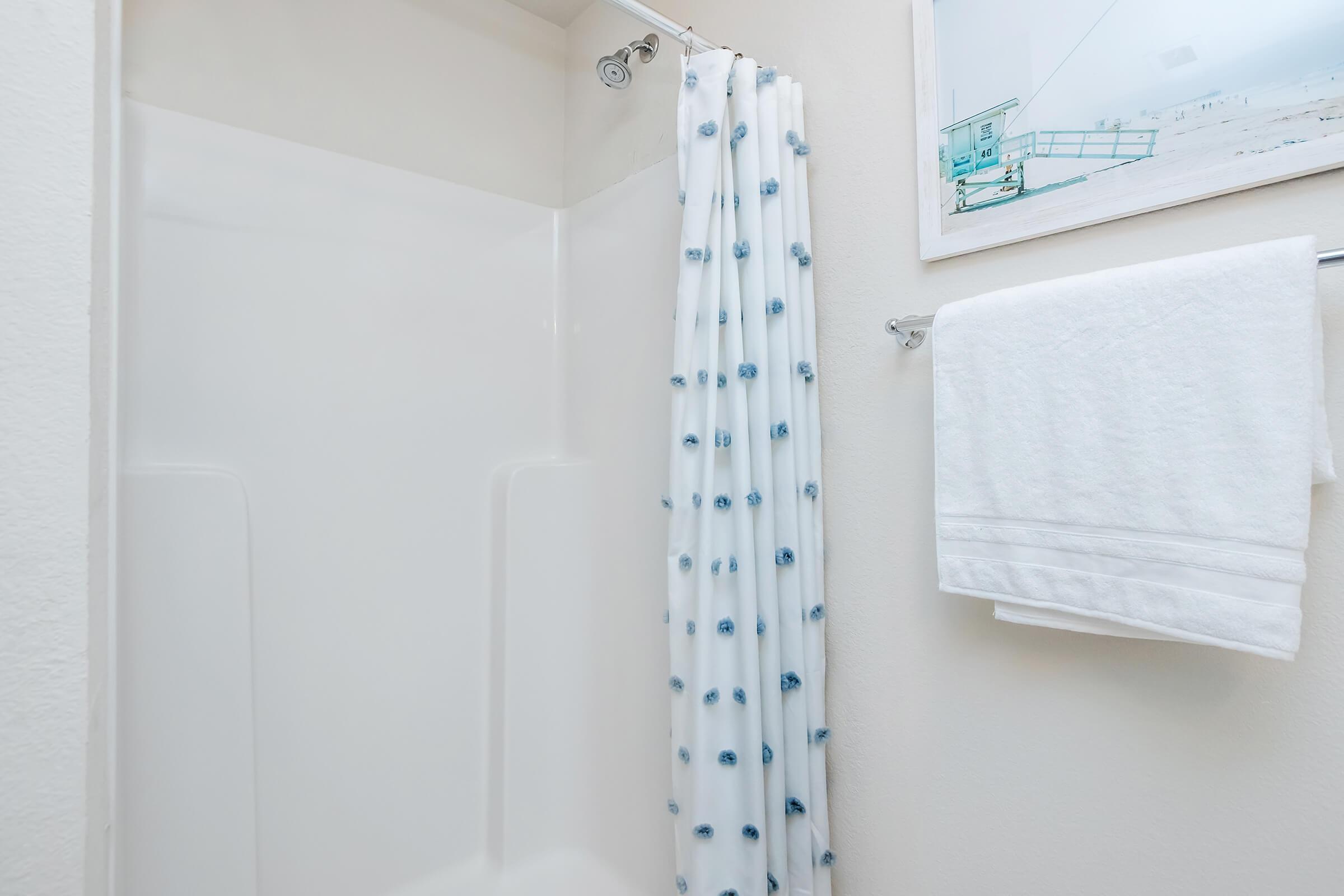 A clean, modern shower space featuring a white shower curtain with blue accents, a neatly hung white towel on a rail, and a simple, minimalistic design. A framed coastal-themed picture is visible on the wall, adding a touch of decor to the otherwise plain bathroom.