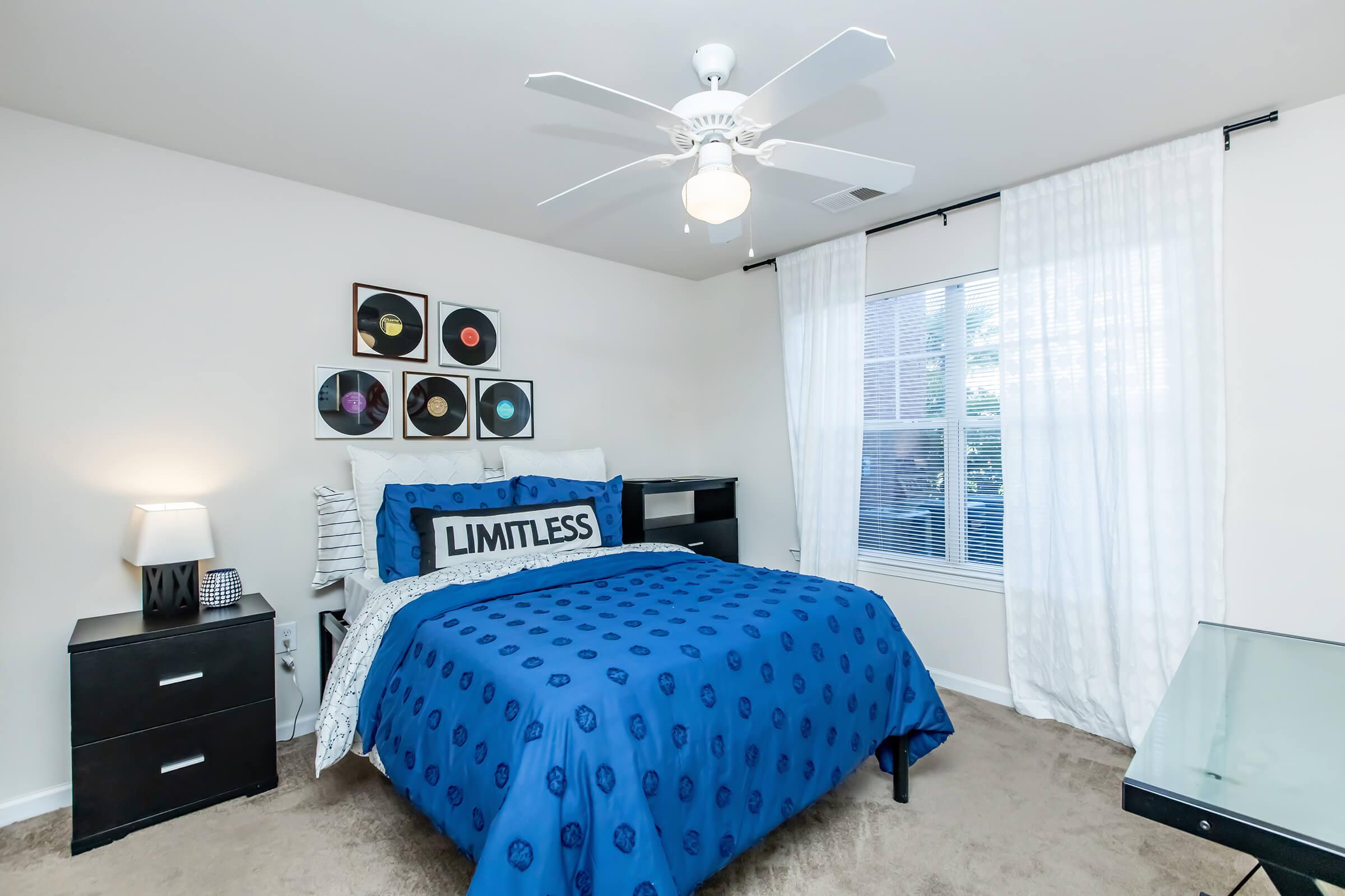 A cozy bedroom featuring a blue bedspread with "LIMITLESS" text, decorative vinyl records on the wall, a bedside lamp, and a black nightstand. Natural light comes through a window with sheer curtains, and a ceiling fan adds a modern touch. The room has beige carpet and a sleek desk.