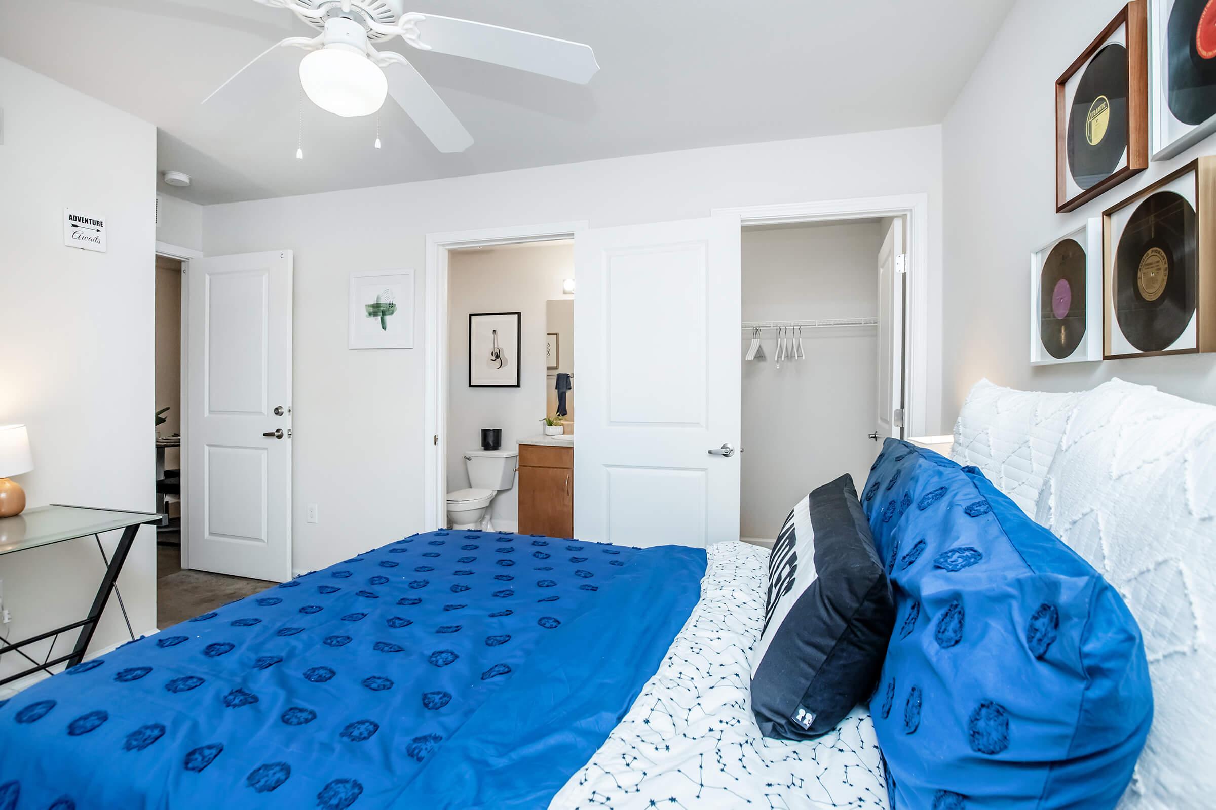 A cozy bedroom featuring a large bed with blue bedding, a white quilt, and decorative pillows. There's a stylish ceiling fan, a glass side table with a lamp, and framed records on the wall. An open doorway leads to a bathroom, and another door is visible in the background. The walls are painted white, creating a bright atmosphere.