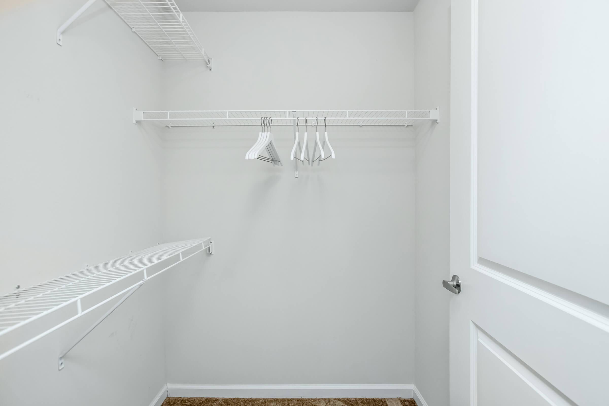 An empty closet with white wire shelving on two walls. Several empty hangers are hanging from one shelf, while the other shelf is also empty. The walls are painted a light gray color, and the floor is covered with a brown carpet. A white door is partially visible on the right side.