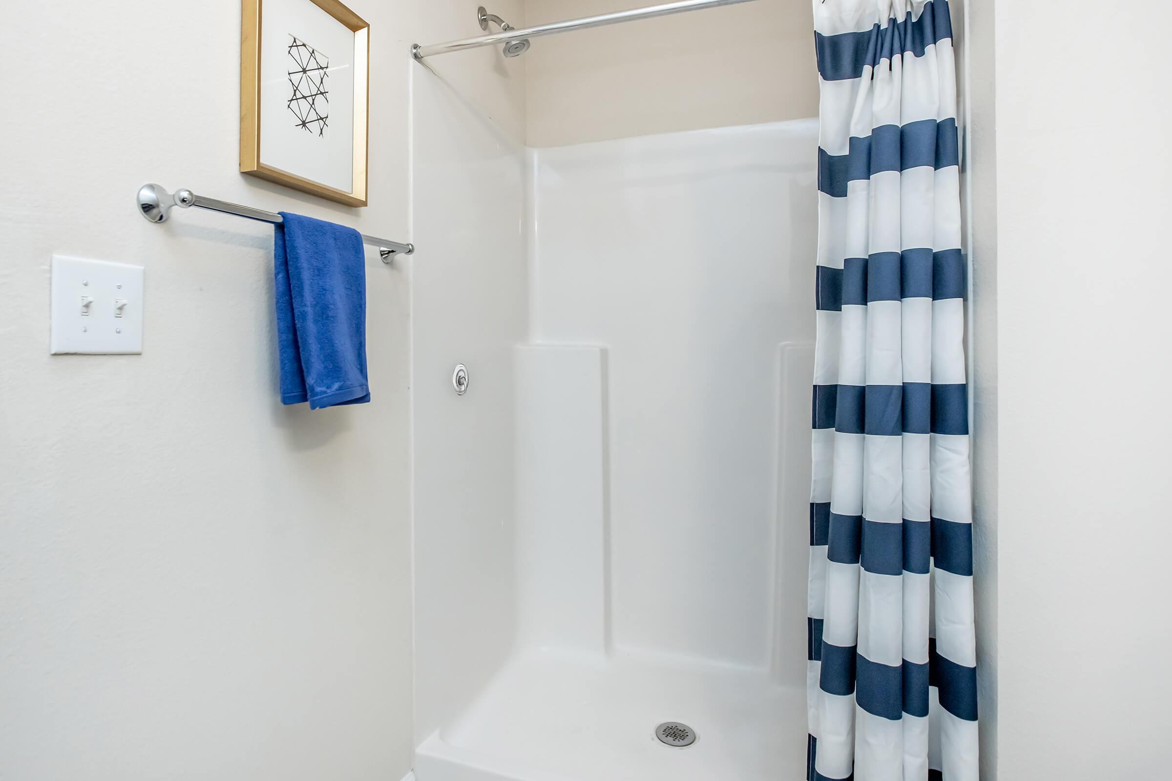 A clean bathroom shower area featuring a white shower stall, a navy and white striped shower curtain, a blue towel hanging on a towel rack, and a decorative framed artwork on the wall. The overall look is modern and tidy, with neutral wall colors.
