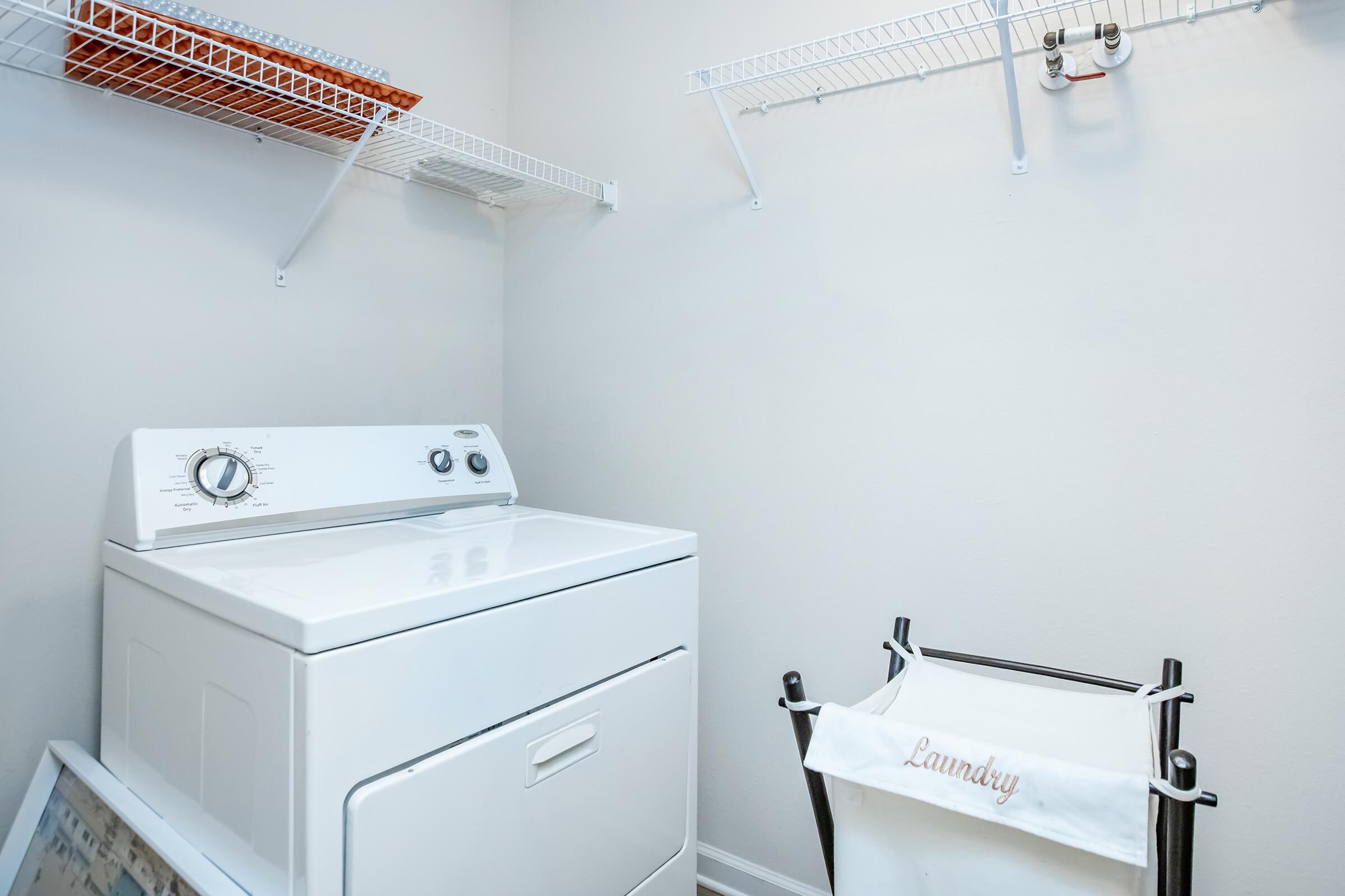 A clean laundry room featuring a white top-loading washing machine, a laundry basket on a shelf, and a black laundry cart with a white fabric liner labeled "Laundry." The walls are painted light colors, and there are wire shelves for storage.