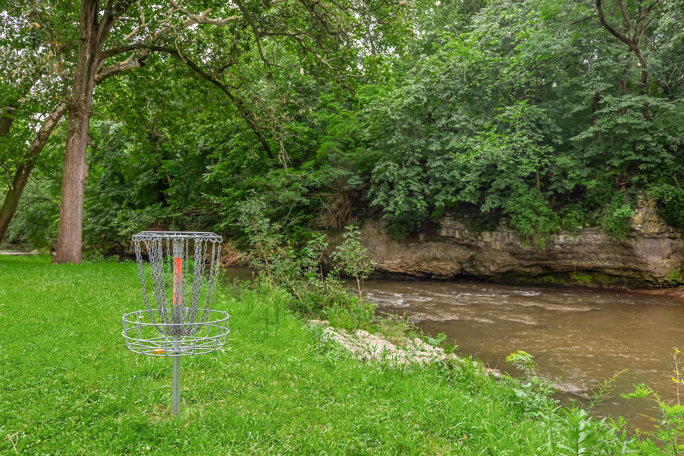 A disc golf basket sits on a grassy area next to a winding creek, surrounded by lush green trees and rocky terrain. The tranquil scene features a mix of natural elements, with the water reflecting the overhanging foliage, creating a peaceful outdoor setting.