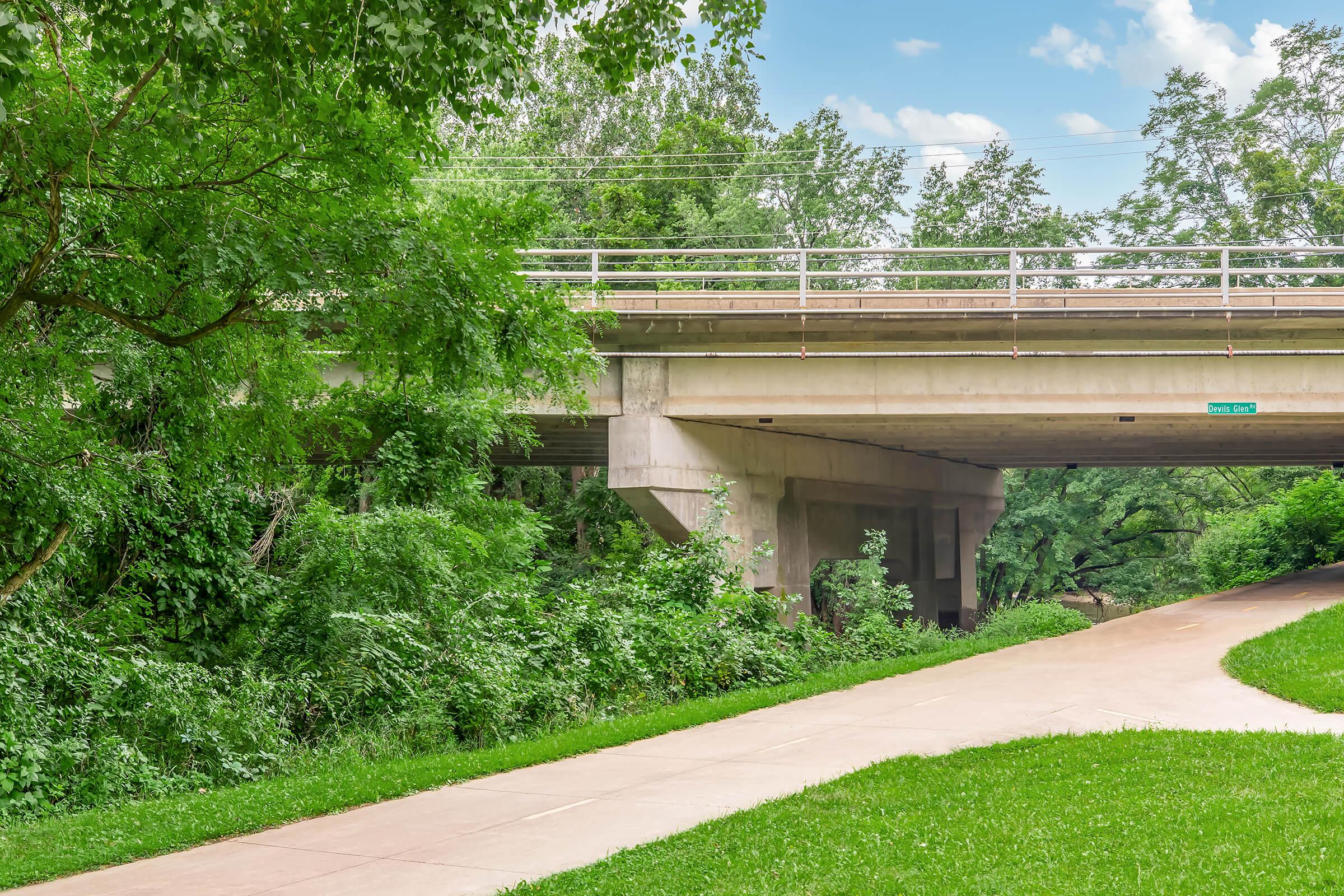 A concrete bridge spans over a lush green area, with trees and vegetation surrounding it. Below the bridge, a paved path winds through the greenery, inviting pedestrians or cyclists to explore the landscape. The sky is partly cloudy, adding a serene atmosphere to the scene.
