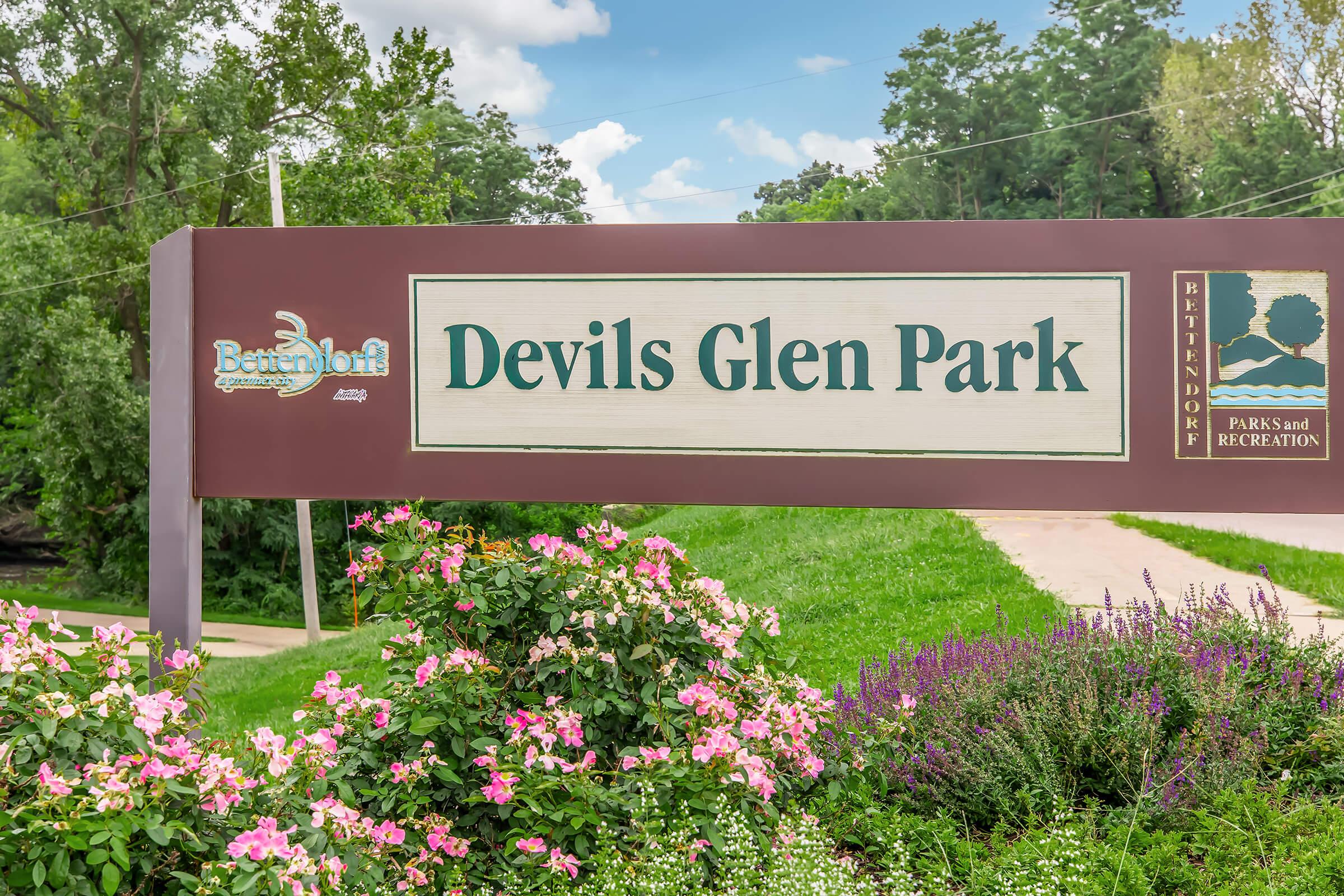 Sign for Devils Glen Park, framed by colorful flowers, with a green landscape and trees in the background. The sign includes the logo for Betterhoff Parks and Recreation, indicating the park's location and offering recreational activities.