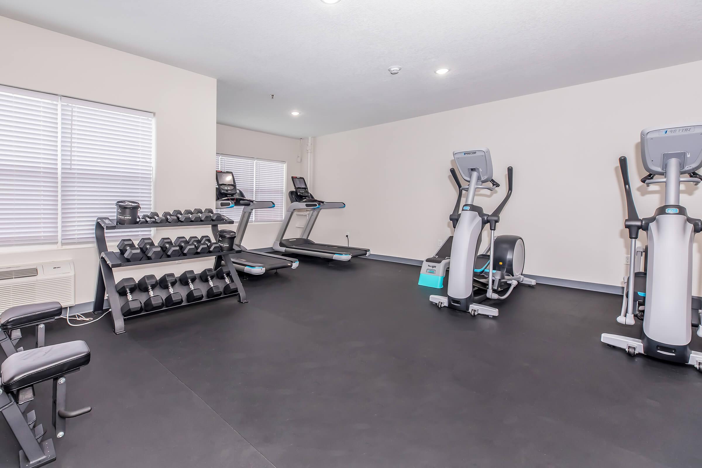 A modern gym interior featuring several pieces of exercise equipment, including ellipticals and a weight rack with dumbbells. The room has large windows allowing natural light, and the flooring is rubberized for safety and comfort.