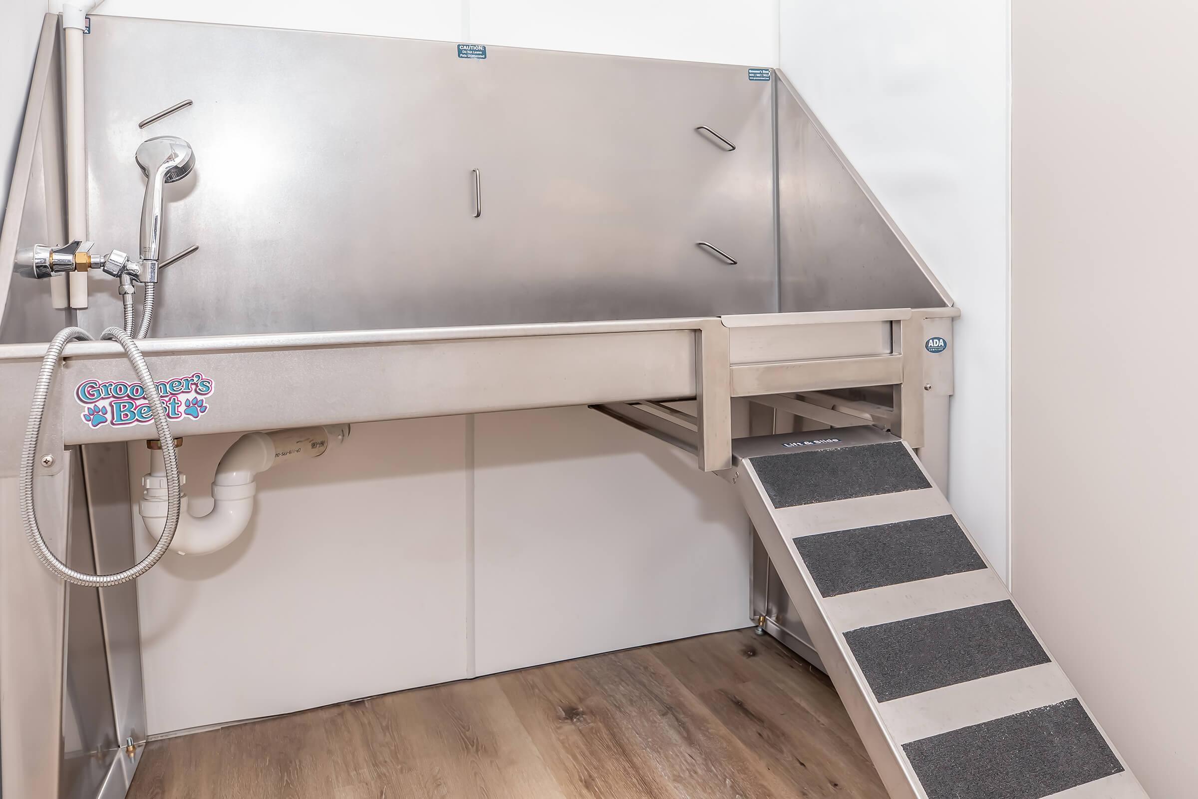 A stainless steel pet bathing station with a ramp leading up to it. The station features a shower fixture on the side and has a clean, minimalist design. The floor is wooden, and the walls are white, creating a bright and functional pet grooming area.