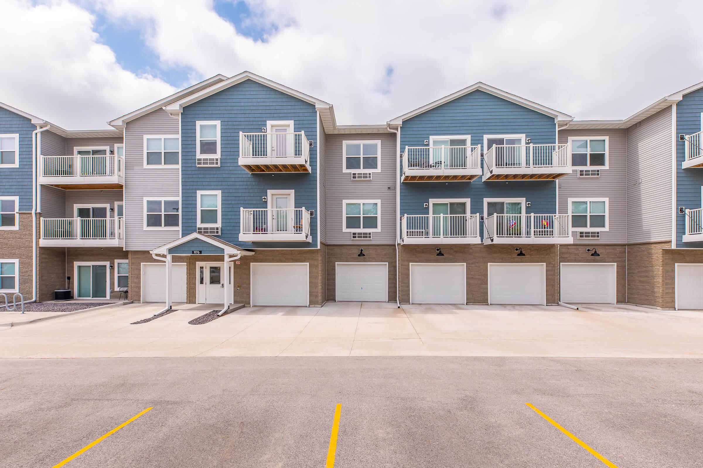 A modern apartment building with blue and gray siding, featuring multiple balconies and garage spaces below. The structure is two stories tall, with a clear sky and scattered clouds in the background. The parking lot has several marked spaces.