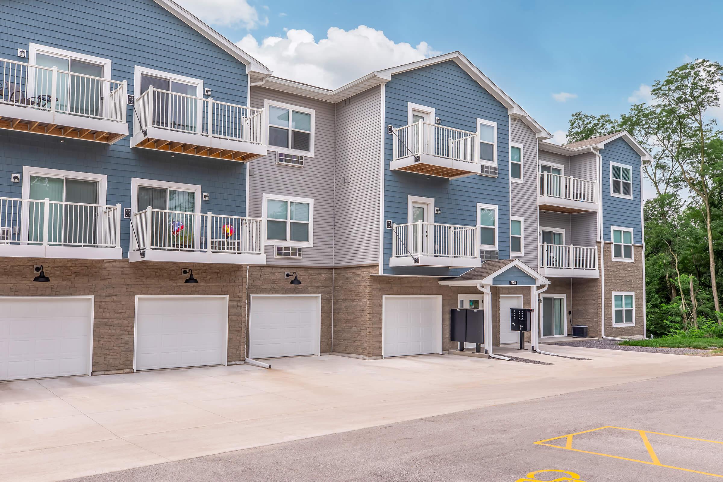Modern apartment building with multiple levels and balconies. The exterior features a combination of gray siding and stone accents. Several garages are visible at the ground level, along with a mail station. The setting includes trees in the background and a clear blue sky.