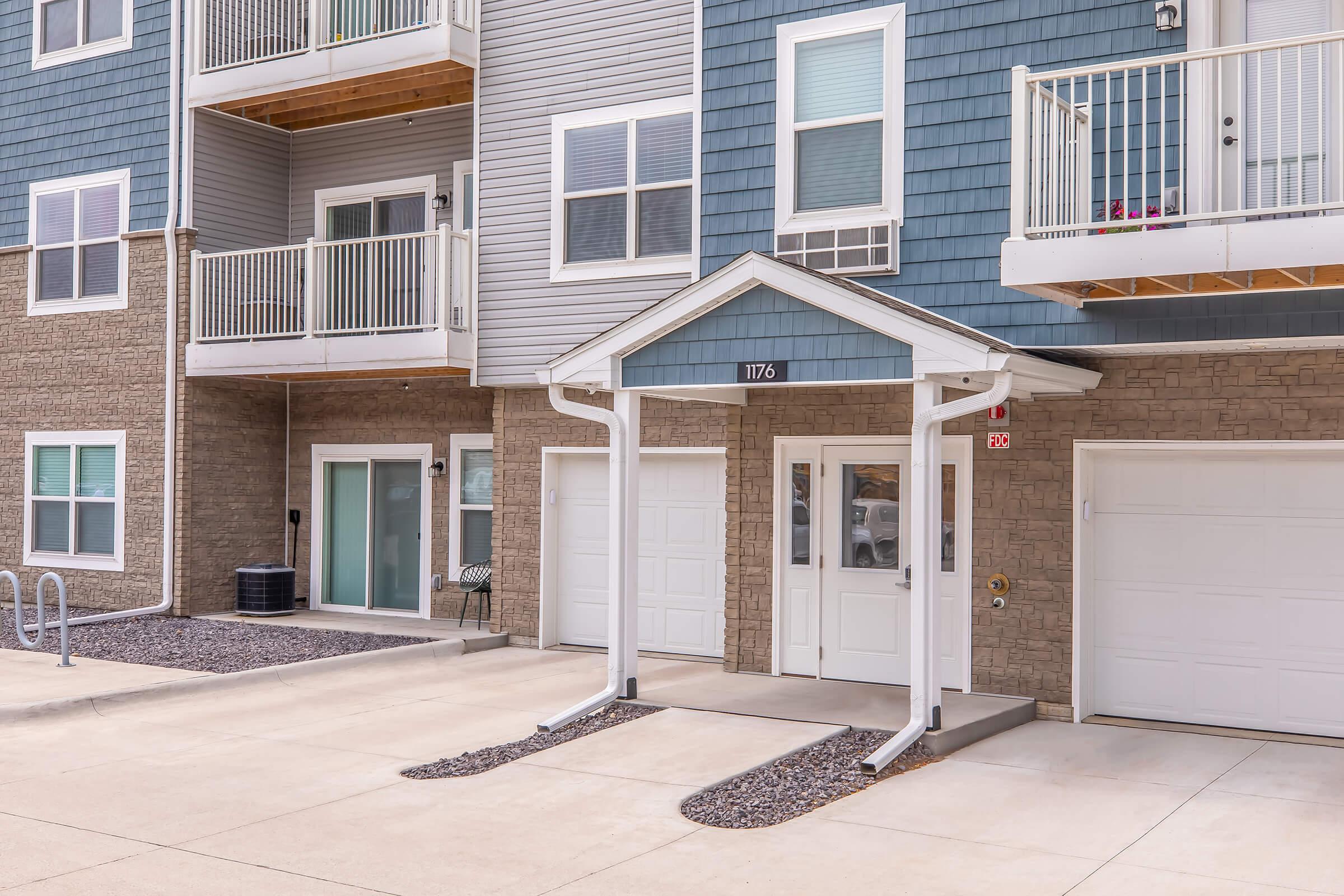 Exterior view of a multi-unit residential building featuring a covered entrance with the number "1176," balconies, and garages. The façade includes a mix of gray vinyl siding and stone accents, surrounded by a concrete driveway and landscaped gravel.