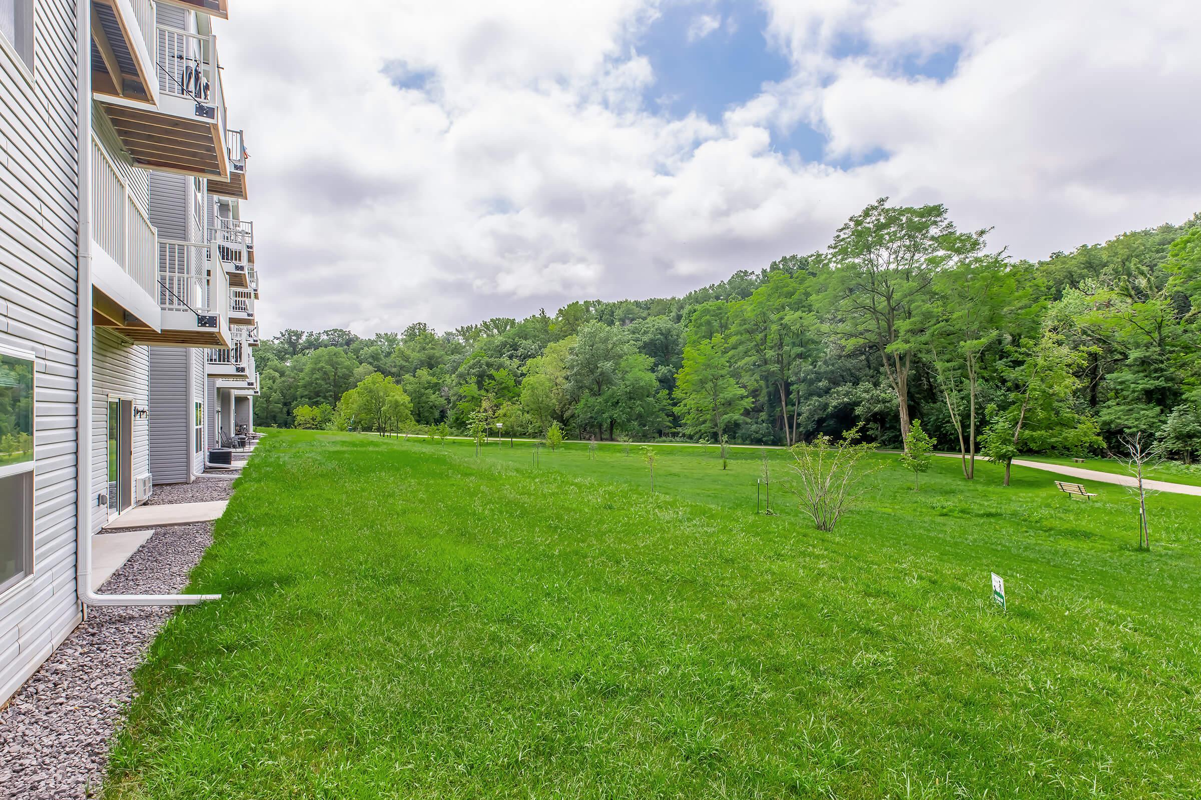 A grassy area beside a residential building, featuring a well-maintained lawn with a few trees and shrubs. In the background, a dense forest is visible under a partly cloudy sky, creating a serene and natural setting.