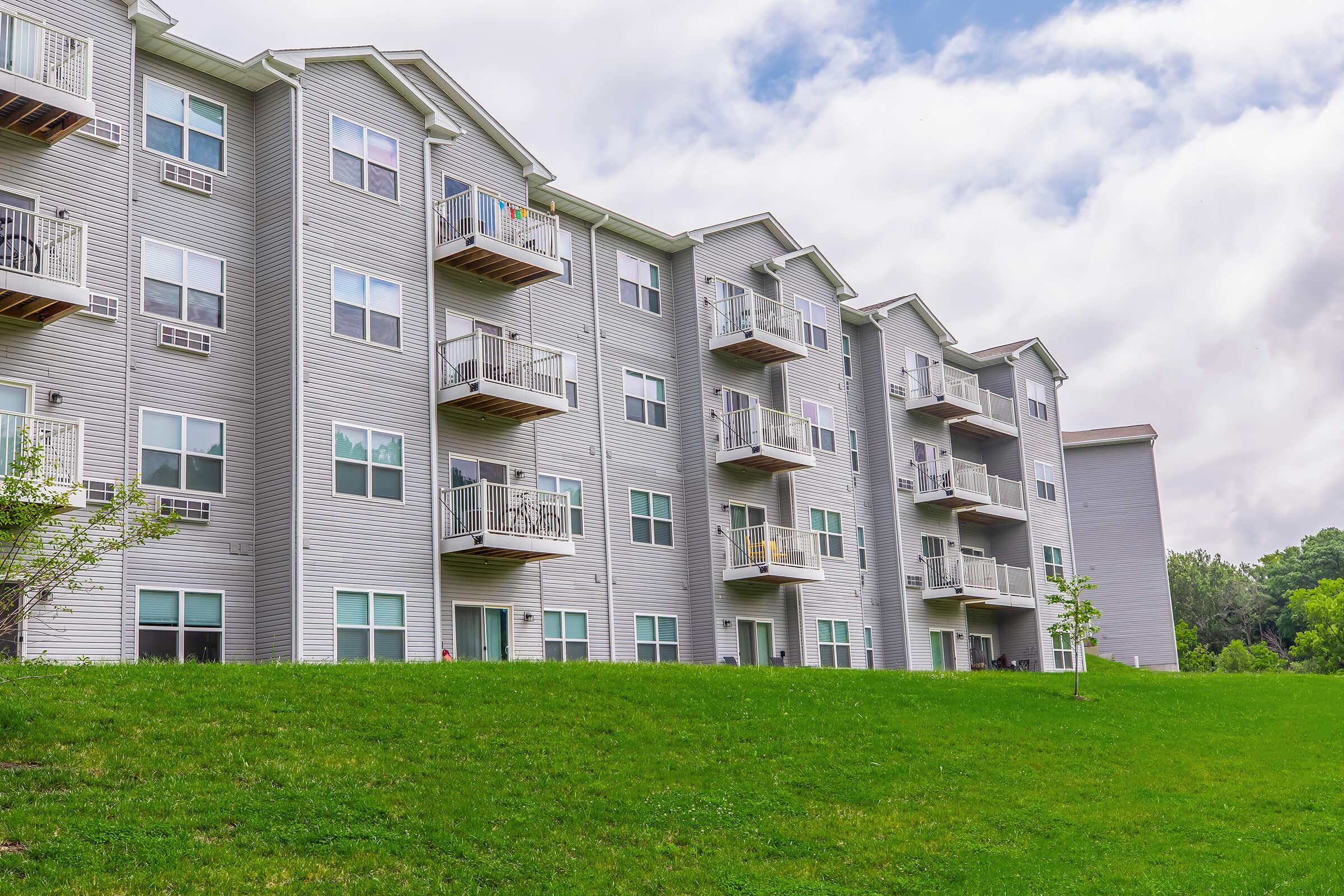 A residential building with multiple gray apartment units, each featuring balconies. The structure is set on a green lawn under a cloudy sky, showcasing a modern design and landscaping with small trees.