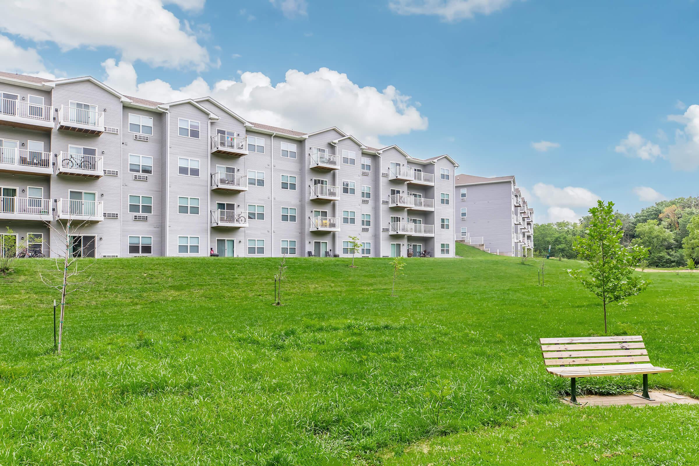A grassy area with a wooden bench in the foreground and several multi-story residential apartment buildings in the background under a blue sky with fluffy clouds. Young trees are scattered throughout the landscape, creating a serene and inviting outdoor space.