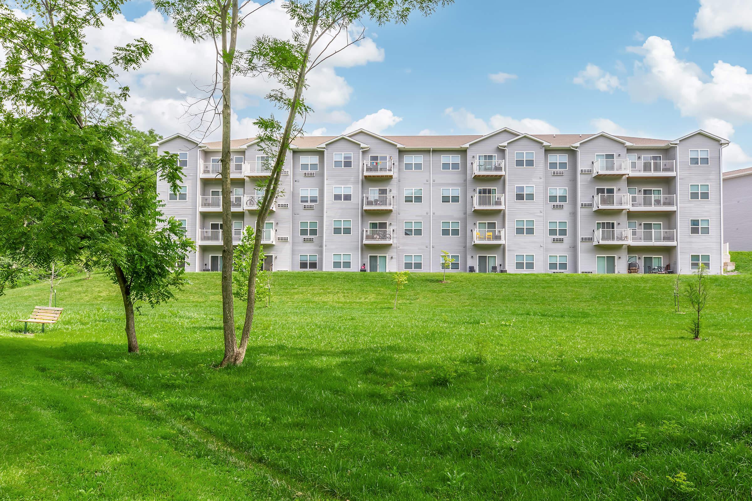A multi-story apartment building with gray siding and multiple balconies, set against a backdrop of a clear blue sky and fluffy clouds. In the foreground, there is a grassy area with sparse trees and a wooden bench, creating a serene and inviting outdoor space.