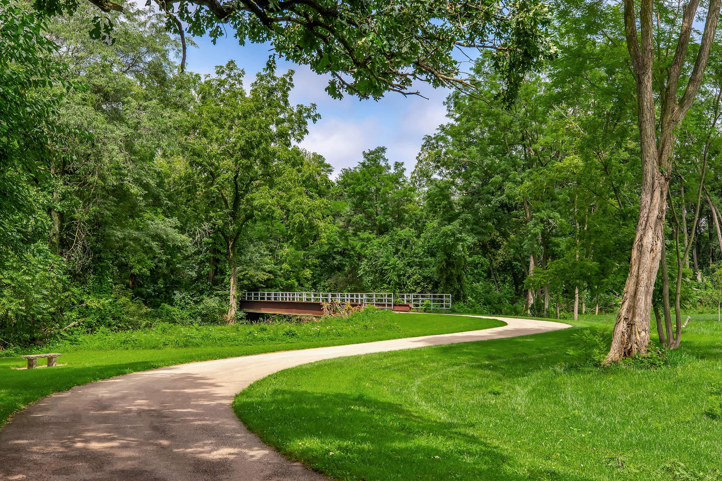 A winding path through a lush green park, with trees lining both sides. In the distance, a wooden bridge crosses a small stream, surrounded by dense foliage. The sky is partly cloudy, providing a serene and tranquil atmosphere. A bench is visible along the path, inviting visitors to relax and enjoy the scenery.