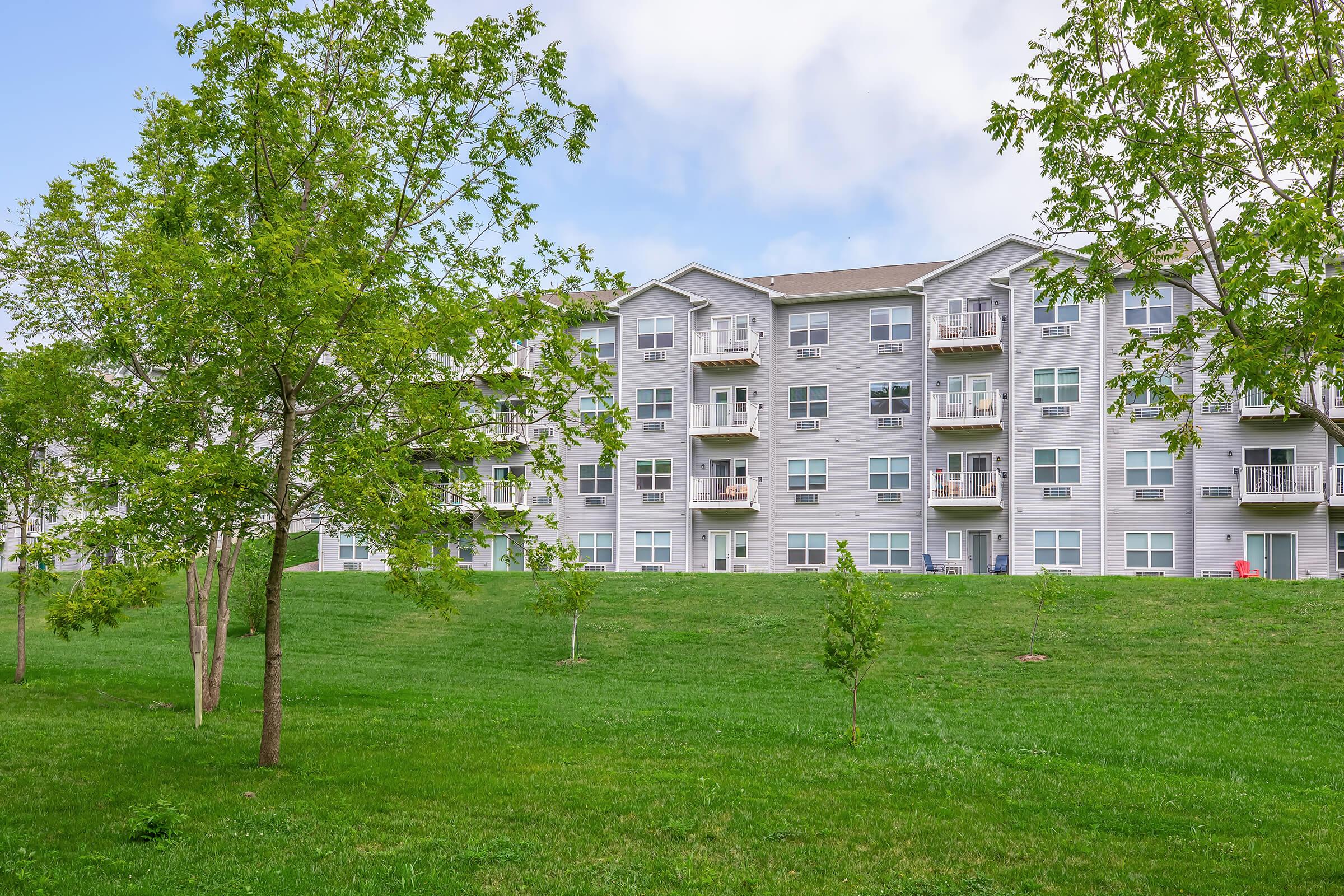 A modern apartment building with multiple balconies, surrounded by lush green grass and young trees. The sky is partly cloudy, creating a serene residential atmosphere.