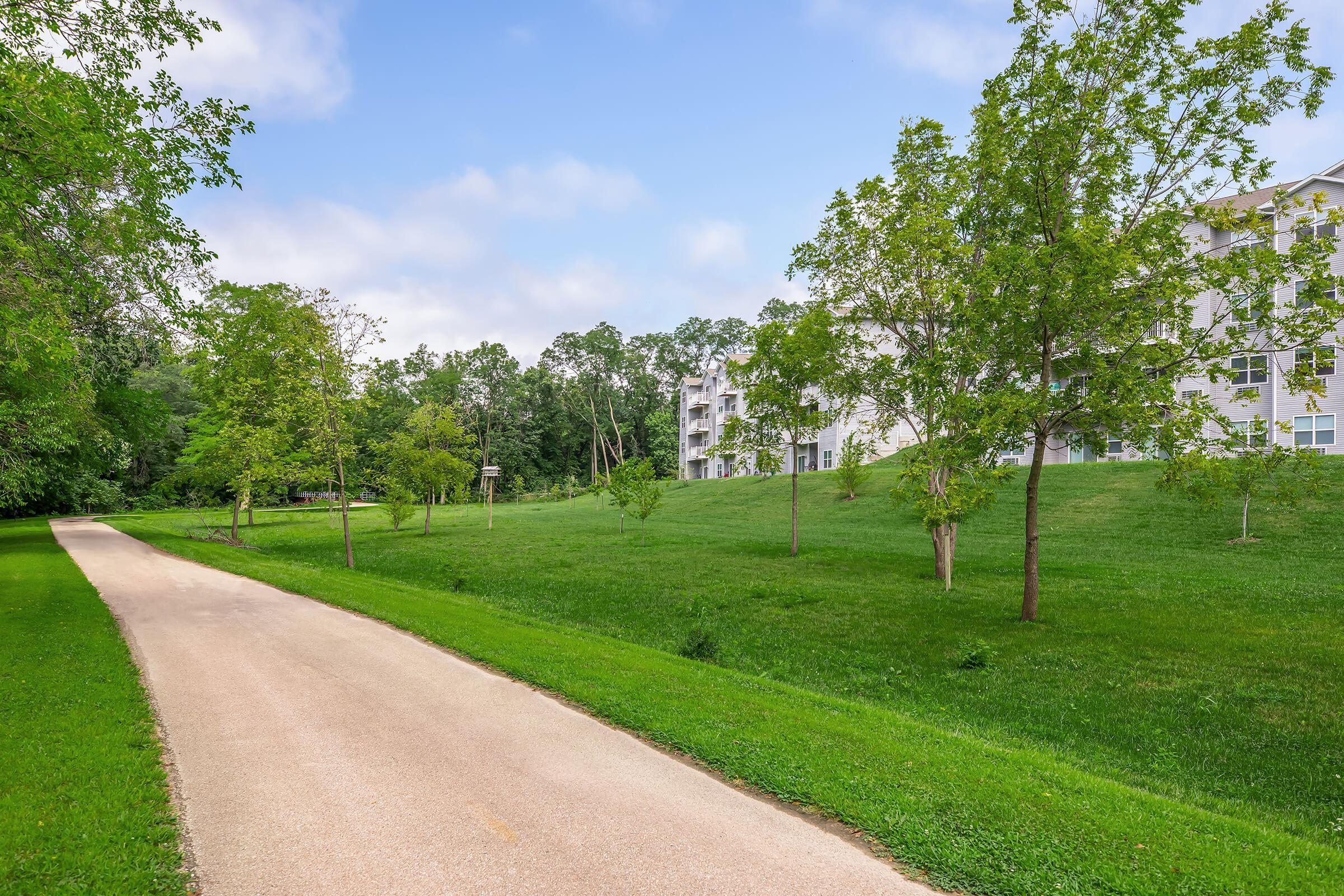A serene pathway bordered by lush green grass and trees, leading through a park-like area. On one side, there are several residential buildings partially visible. The sky is slightly cloudy, creating a calm atmosphere perfect for a leisurely stroll or outdoor activities.