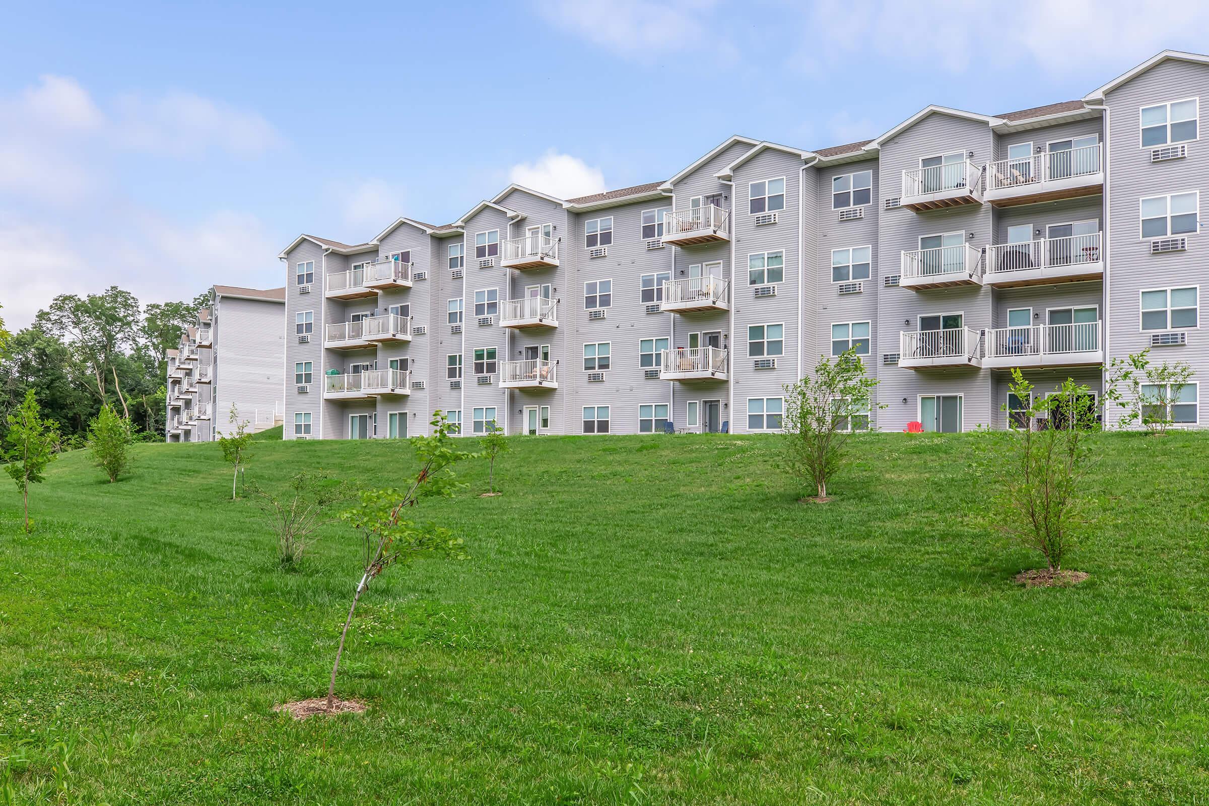 A multi-story residential building with gray siding, featuring multiple balconies. The structure is set on a green lawn with small trees and shrubs in the foreground. The sky is partly cloudy, indicating a pleasant day.