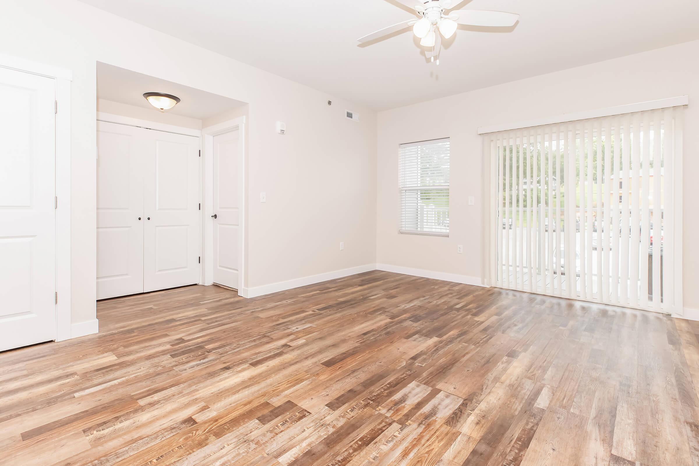 Spacious room featuring light-colored walls, a ceiling fan, and large windows with blinds. The floor has a warm wood finish, and there's a closet to the left. Natural light fills the space, creating an inviting atmosphere. Ideal for living or recreational activities.