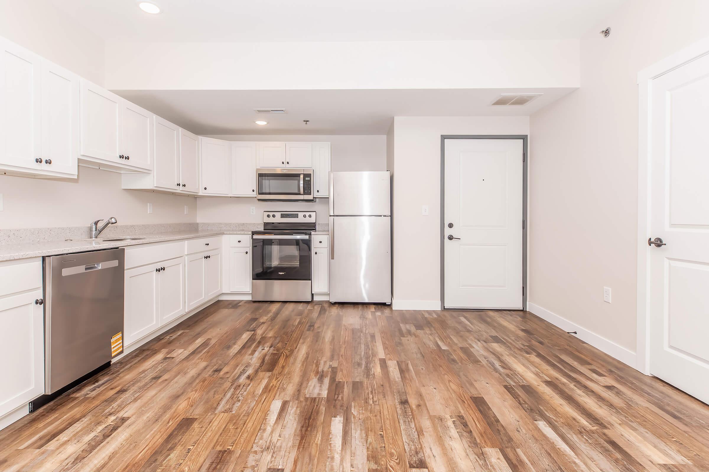 Modern kitchen featuring white cabinetry, stainless steel appliances, and a gray countertop. The space includes a refrigerator, microwave, oven, and dishwasher. The floor is made of wood-like laminate. Natural light brightens the room, which has light walls and a door leading outside.