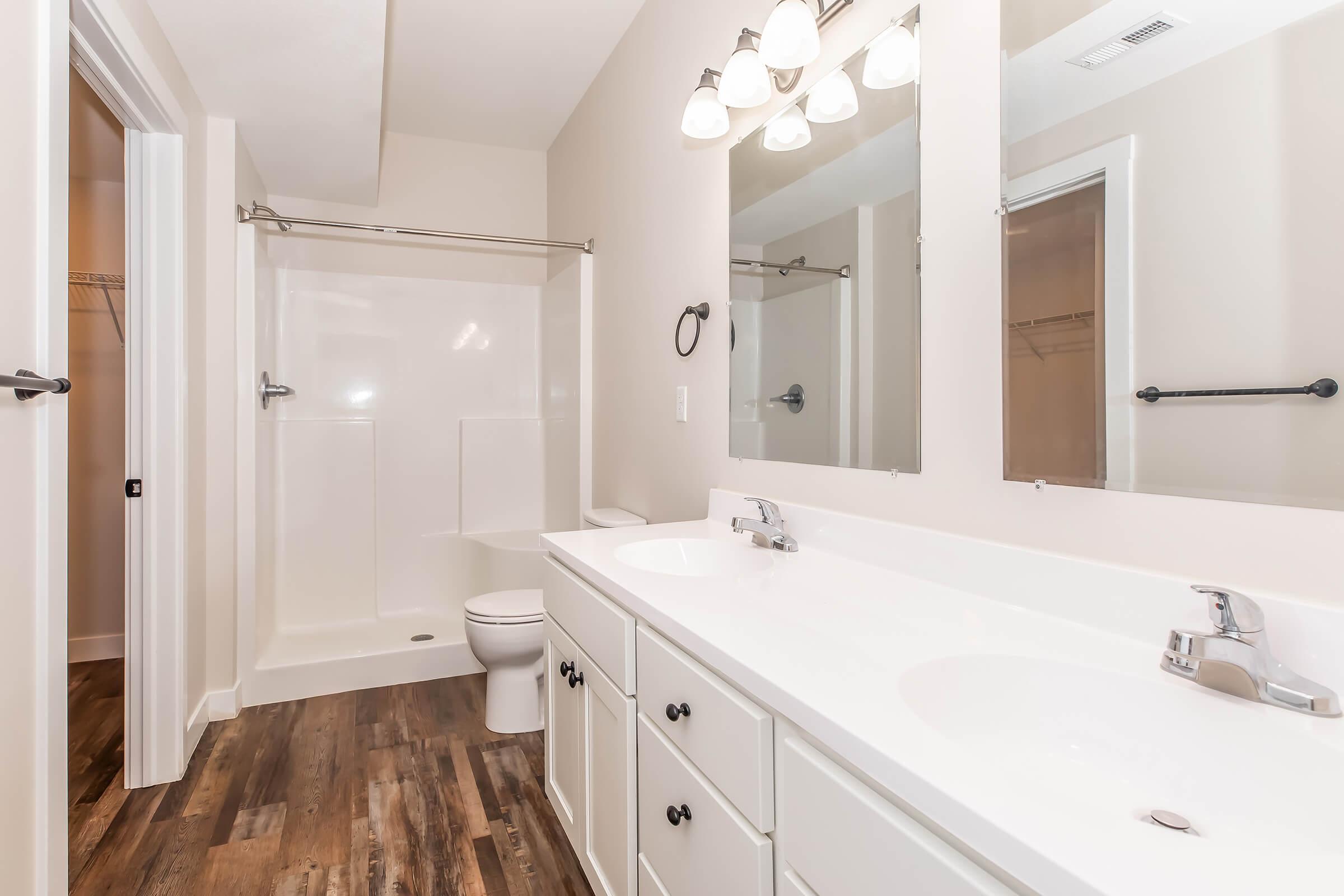 A modern bathroom featuring a double vanity with two sinks, a large mirror, and a spacious shower with a glass door. The floor has a wood-like finish, and there is a toilet in the corner. The walls are painted in a light color, creating a bright and airy atmosphere.